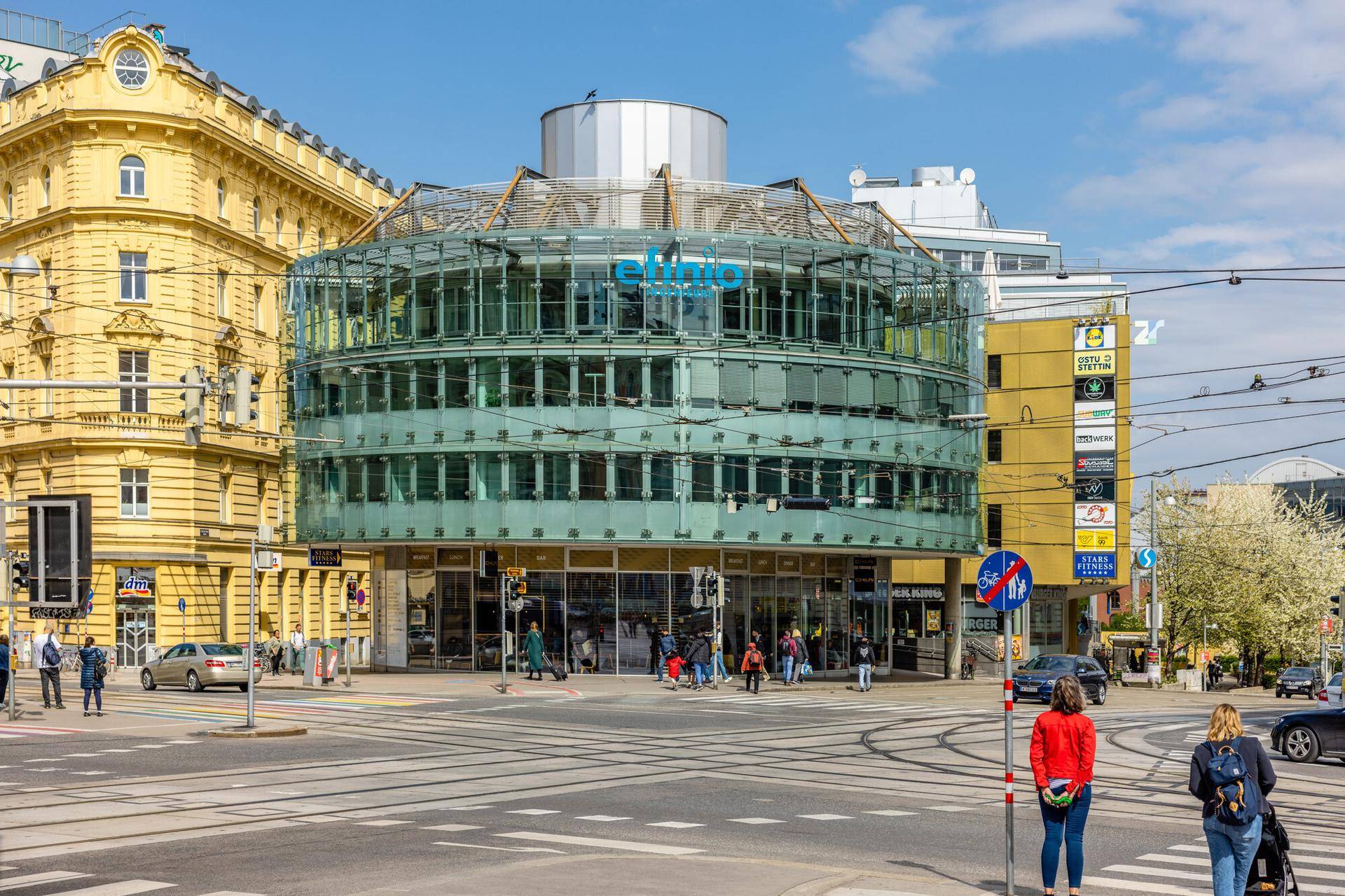 Markantes, rundes Gebäude mit Glasfassade an einer städtischen Kreuzung, umgeben von weiteren Gebäuden.