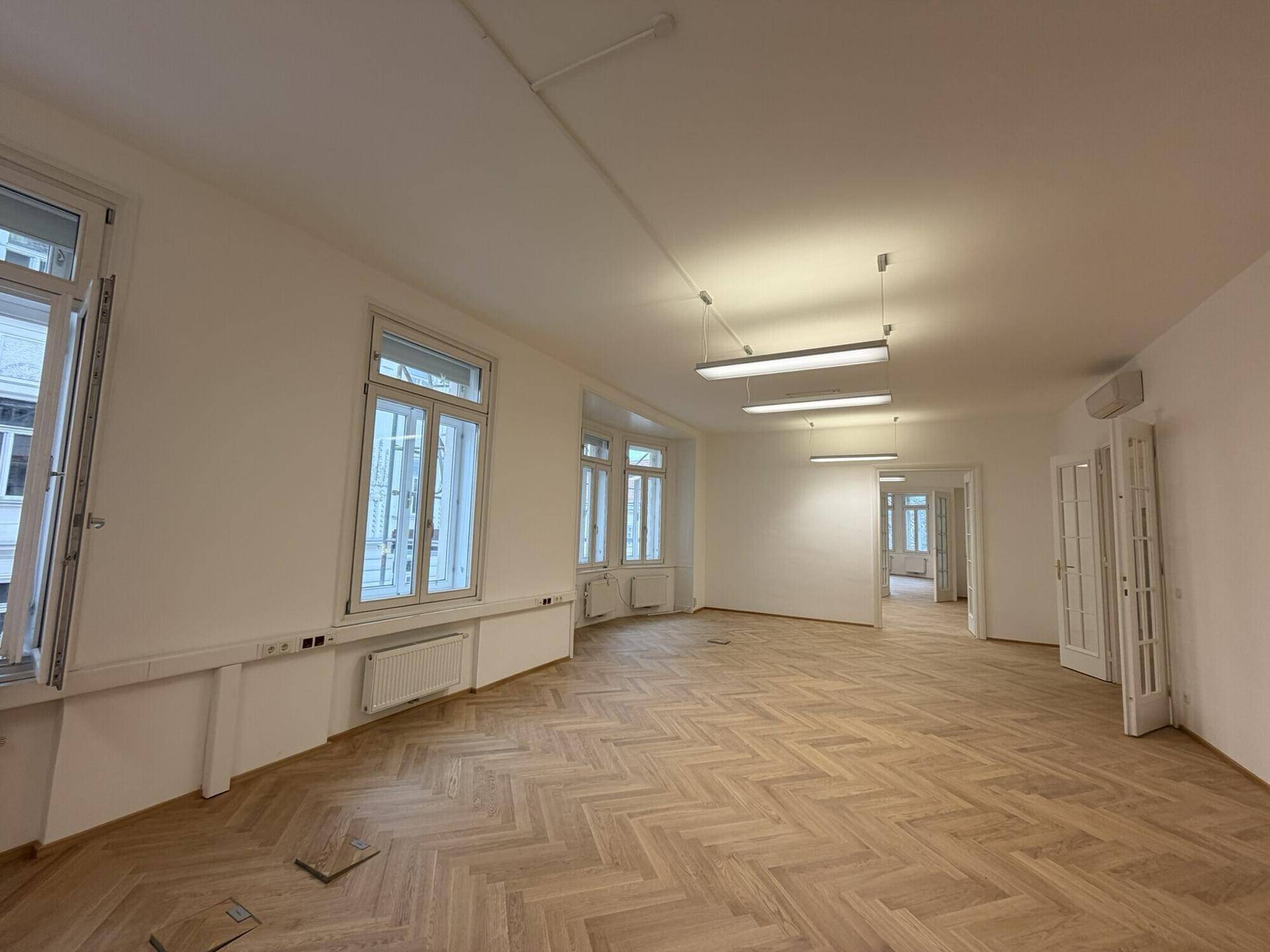 Langer, heller Raum mit Fischgrätparkett und mehreren Fenstern, der in weitere Zimmer führt.