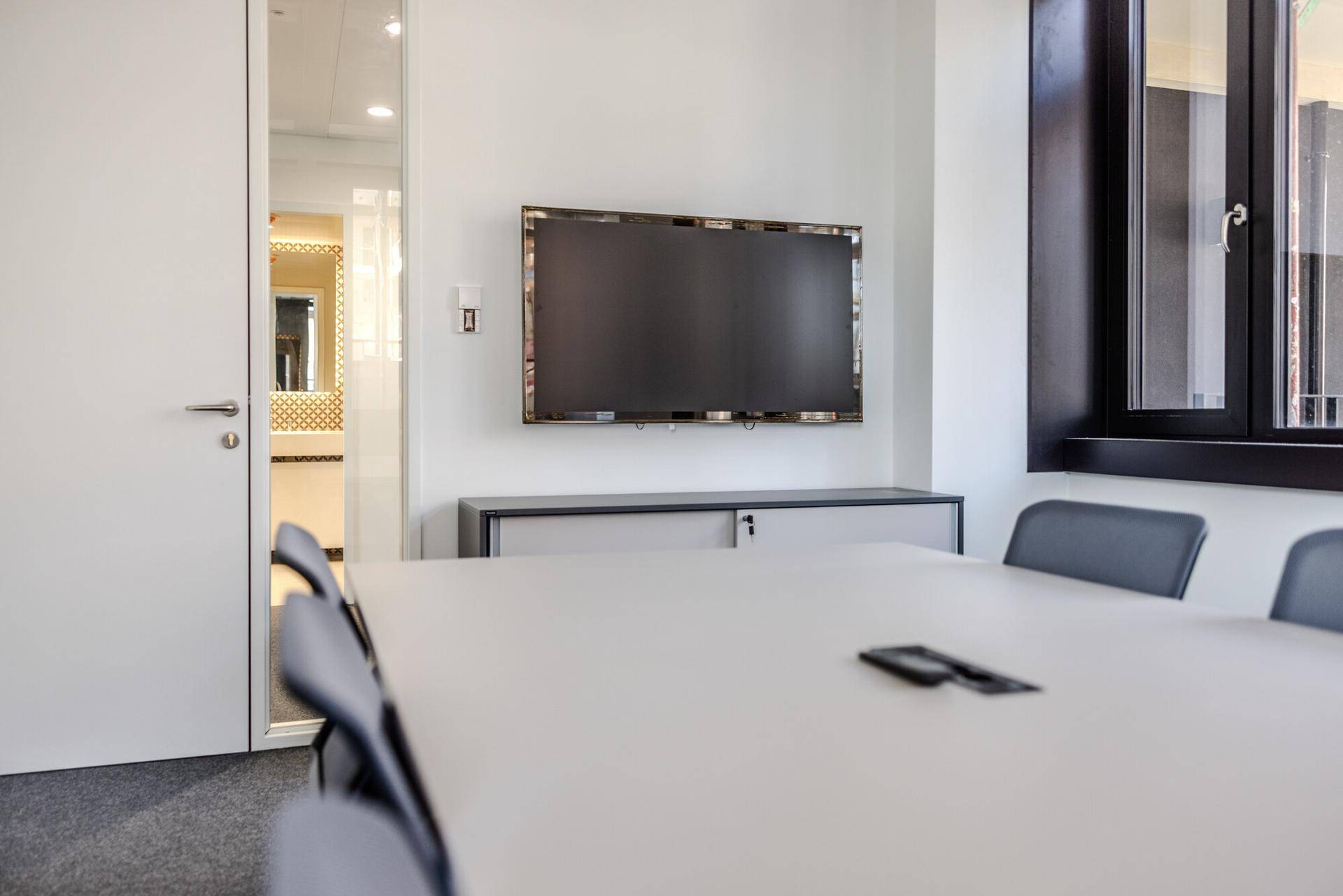 Eleganter Konferenzraum mit großem Tisch, Wandfernseher und Blick auf einen angrenzenden, hellen Waschraum.