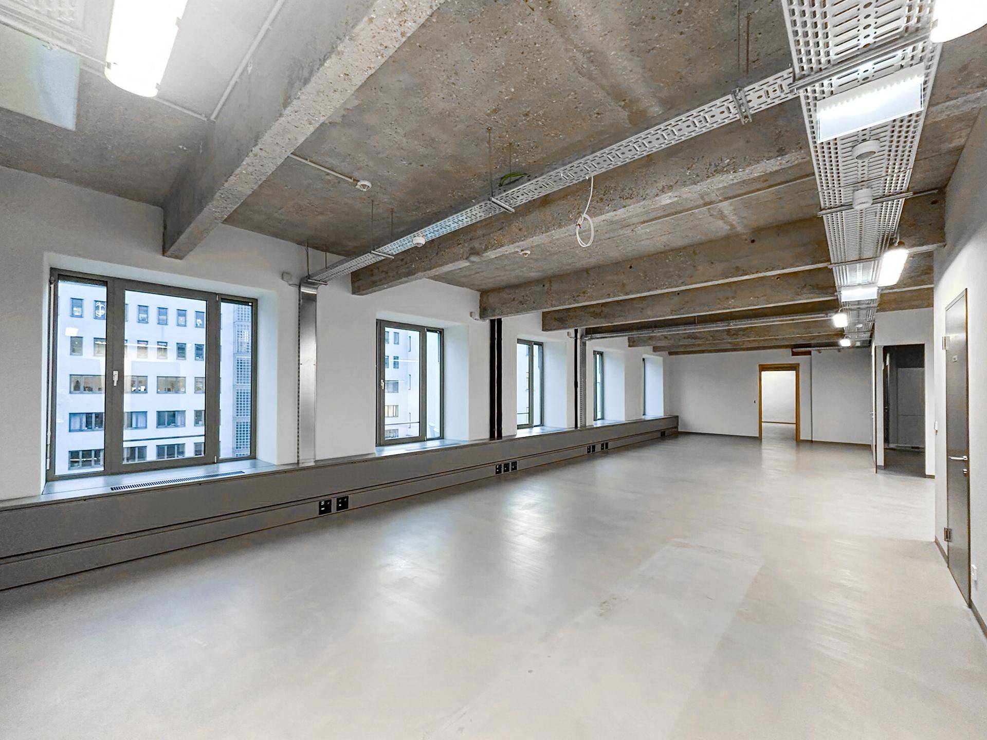 Weitläufiger Bürobereich mit freiliegenden Betondecken und großen Fenstern, die viel Tageslicht hereinlassen.