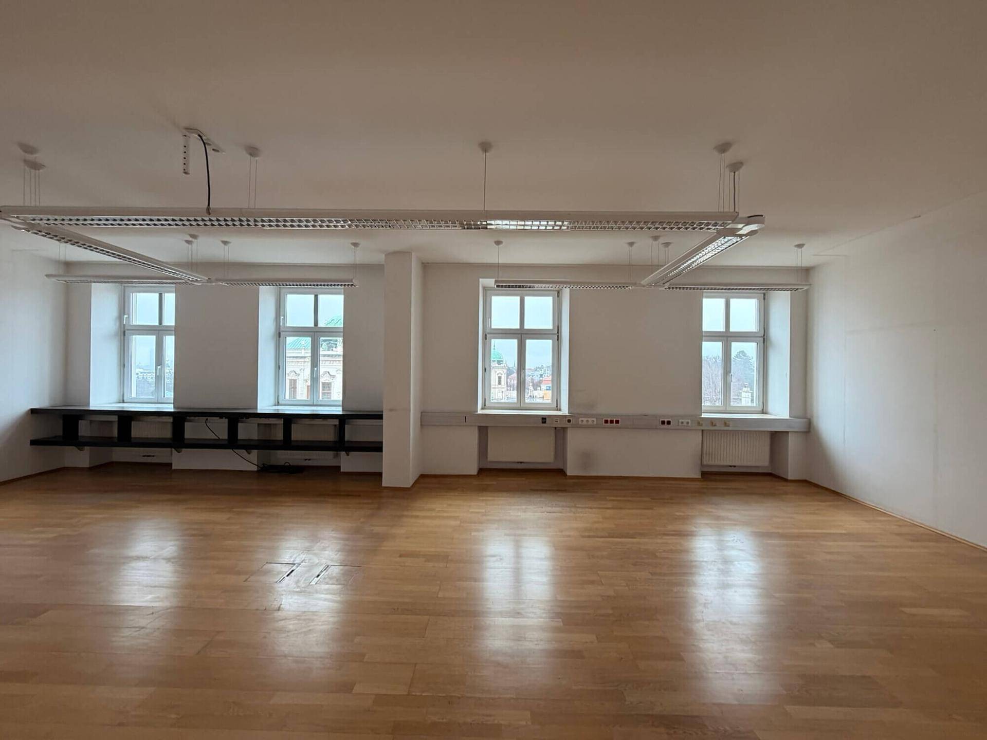 Großer Büroraum mit Holzboden, mehreren Fenstern und langen Leuchtstoffröhren an der Decke für eine helle Arbeitsumgebung.