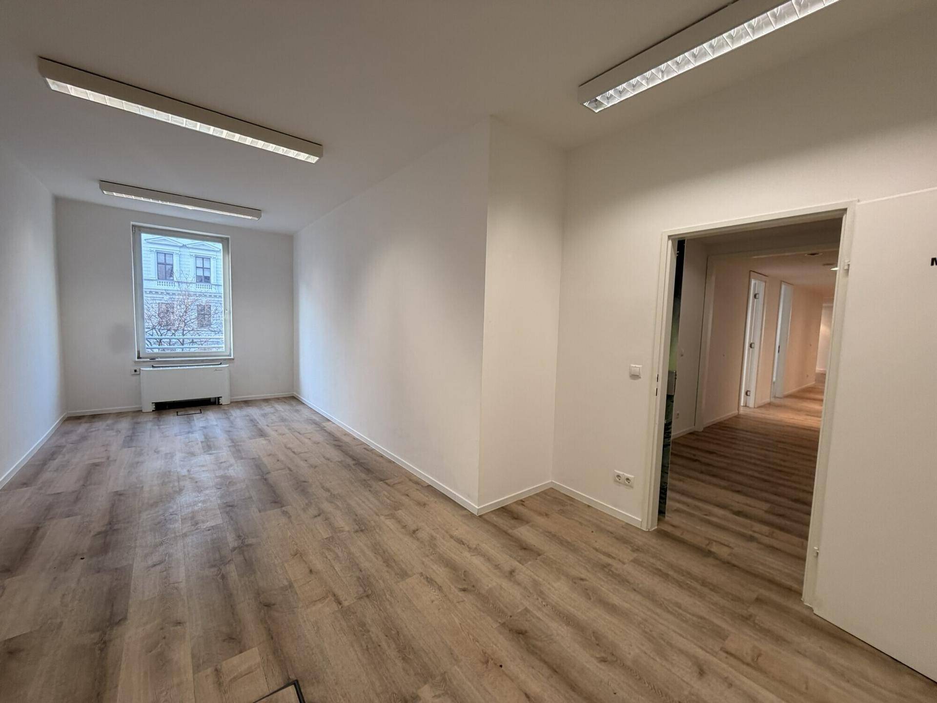 Heller Büroraum mit Laminatboden und einem Fenster, das Tageslicht hereinlässt.