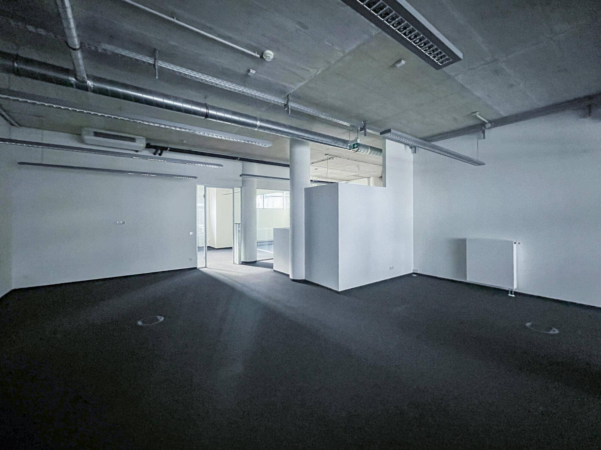 Offener Bürobereich mit Teppichboden, freiliegenden Leitungen und modernen Trennwänden aus Glas.