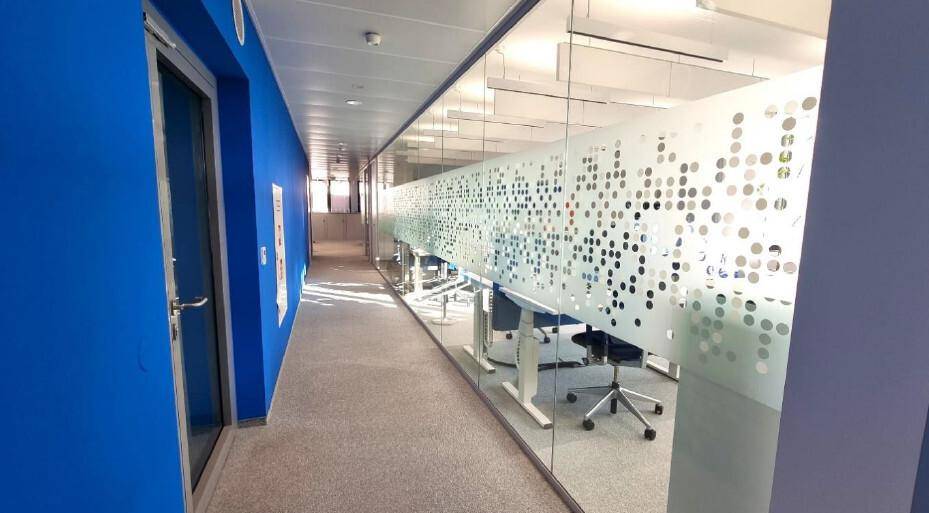 Langer Bürogang mit blauer Wand, Glaswänden zu den Büros und Teppichboden, der eine moderne Ästhetik bietet.