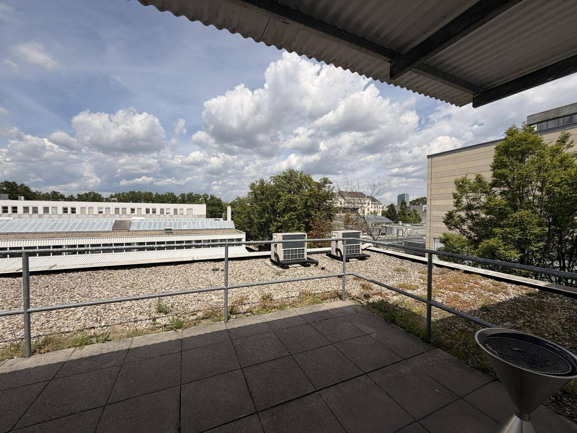 Dachterrasse mit Fliesenboden, Metallgeländer und Blick auf umliegende Gebäude und grüne Bäume unter blauem Himmel.