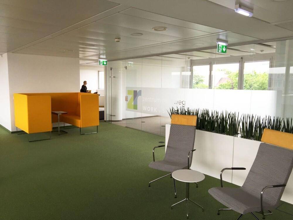 Moderner Loungebereich mit grüner Teppichfläche, bequemen Stühlen und gelben Sitzelementen.