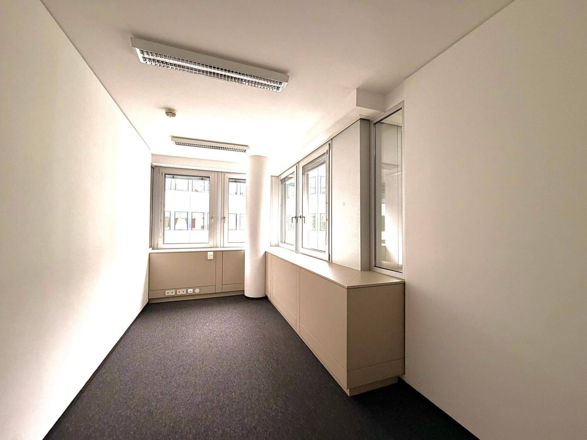 Schmaler Büroraum mit Teppichboden, Fenstern und integrierten Schränken für Stauraum.