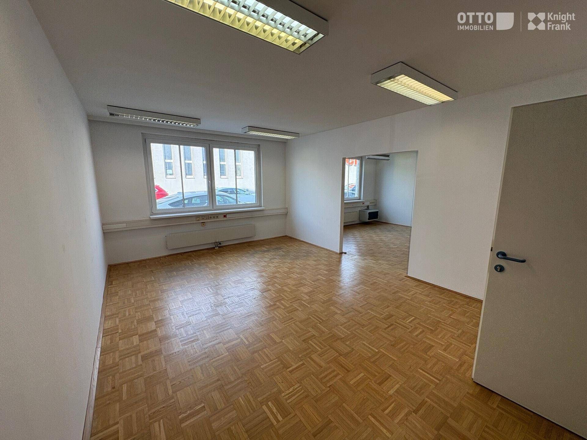 Heller Büroraum mit Parkettboden und Zugang zu einem weiteren Bereich, ideal für flexible Nutzung.
