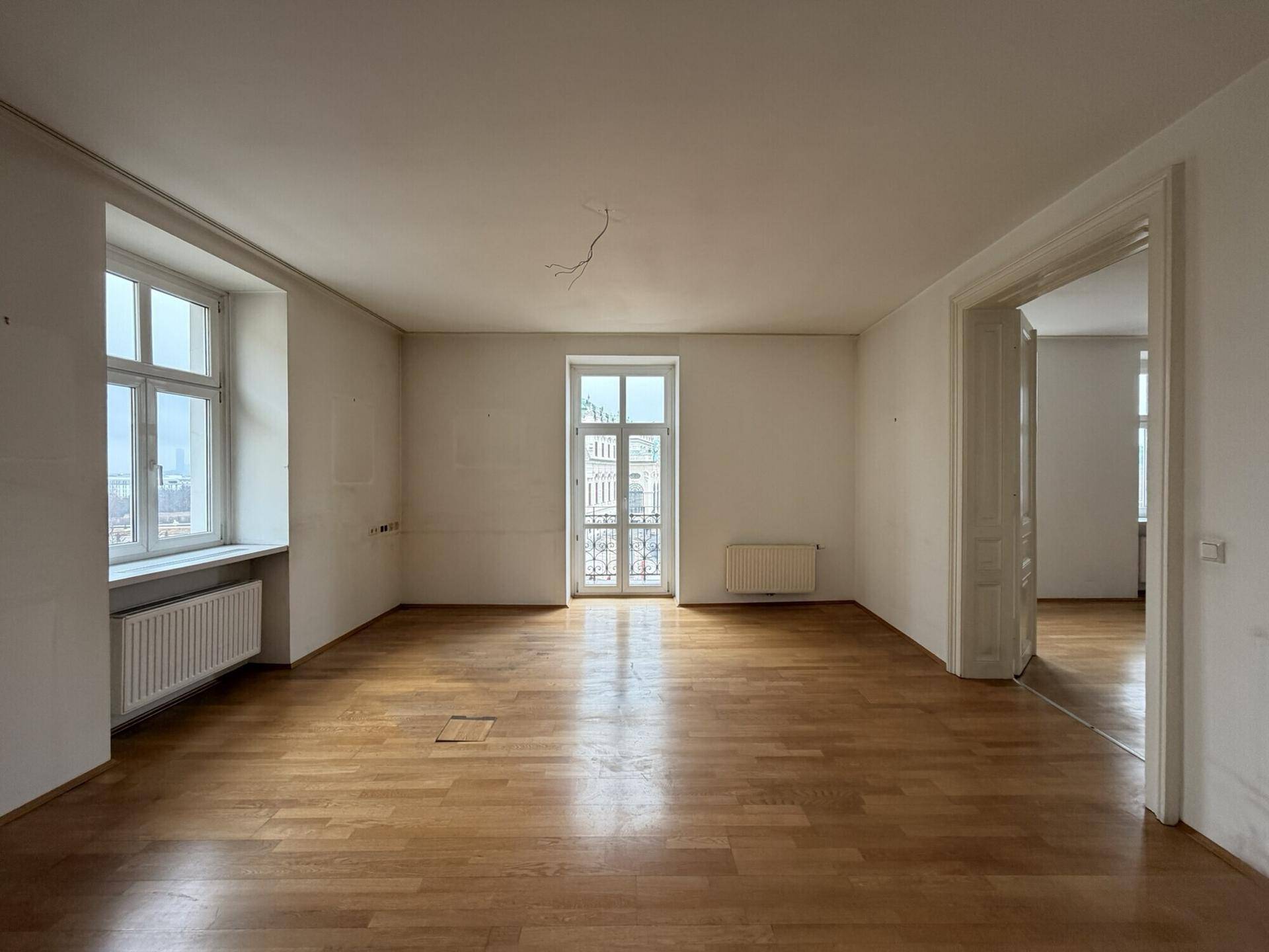 Geräumiger, leerer Raum mit Holzboden, hohen Fenstern und einem französischen Balkon, der viel Tageslicht bietet.