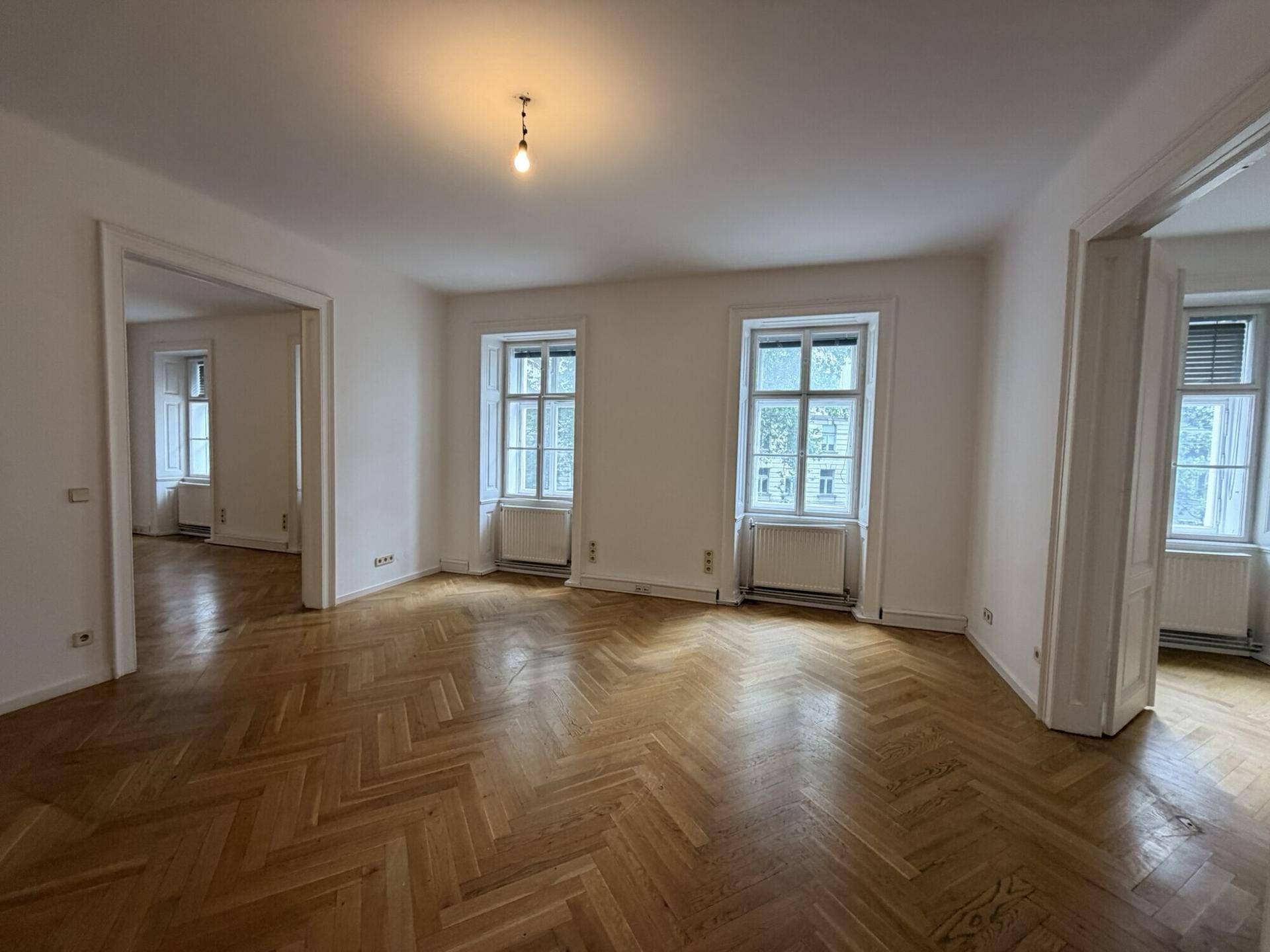 Großes Wohnzimmer mit Fischgrätparkett, hohen Fenstern und Blick in angrenzende Räume.