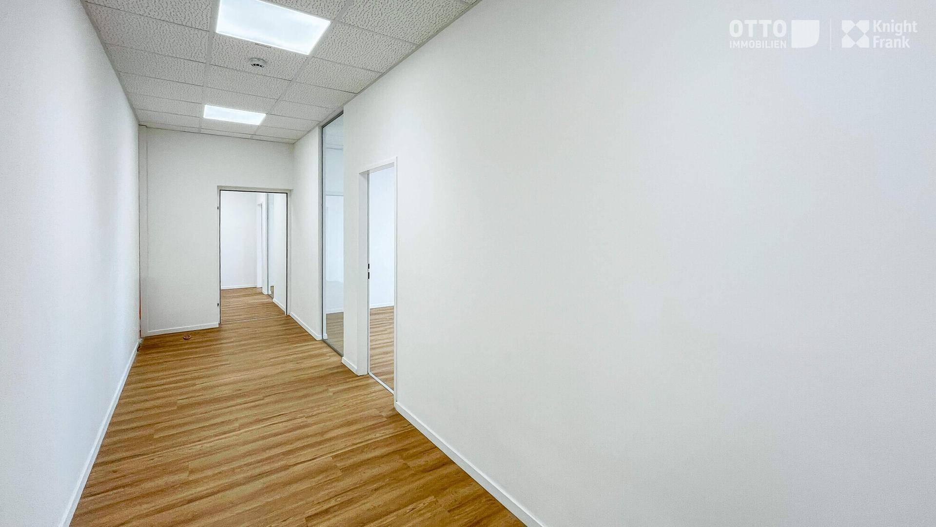 Langer, heller Flur mit Laminatboden und Zugang zu mehreren Büroräumen durch Glastüren.