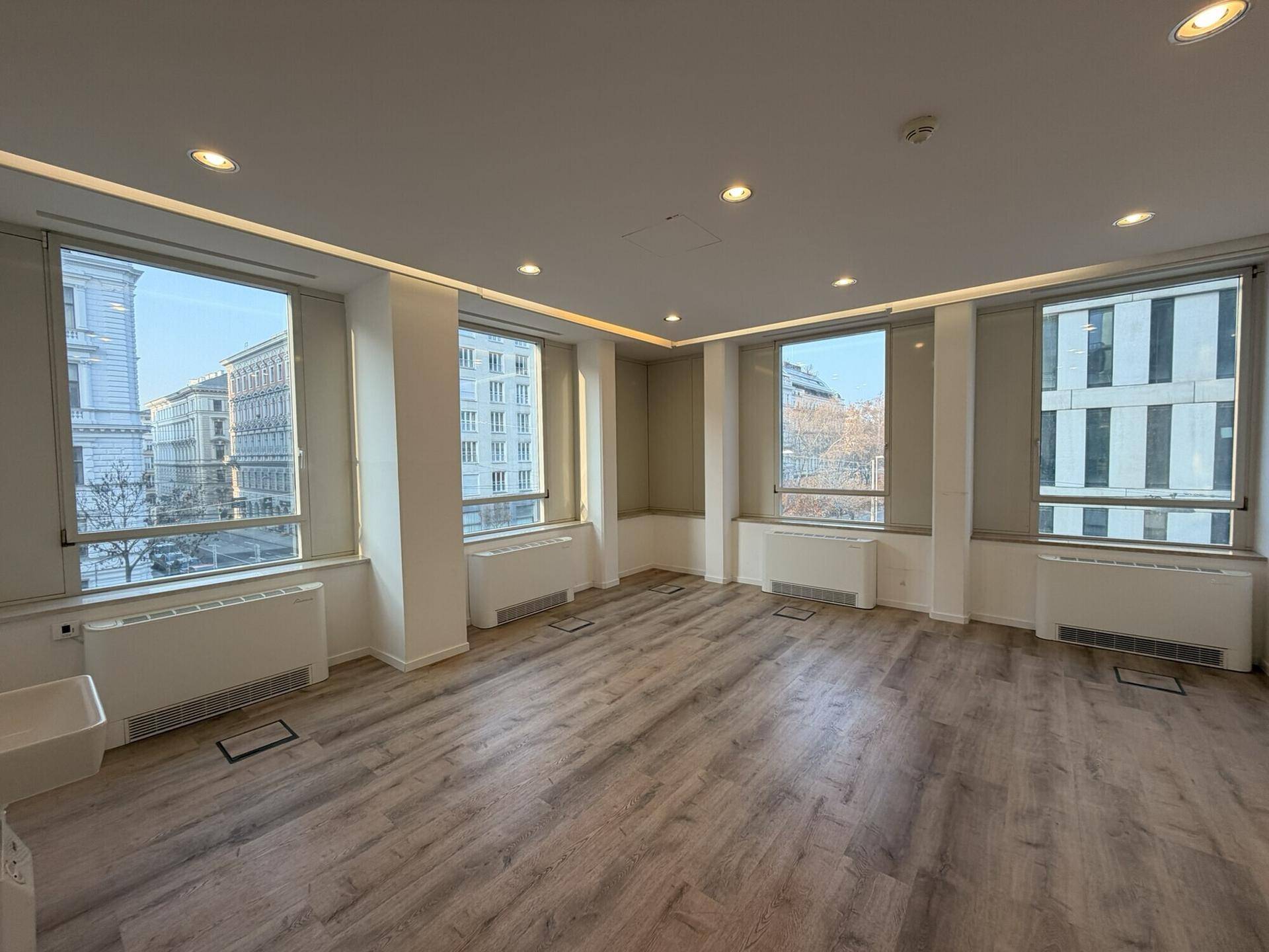 Heller Büroraum mit mehreren Fenstern, die einen Blick auf die Stadt und die umliegenden Gebäude ermöglichen.