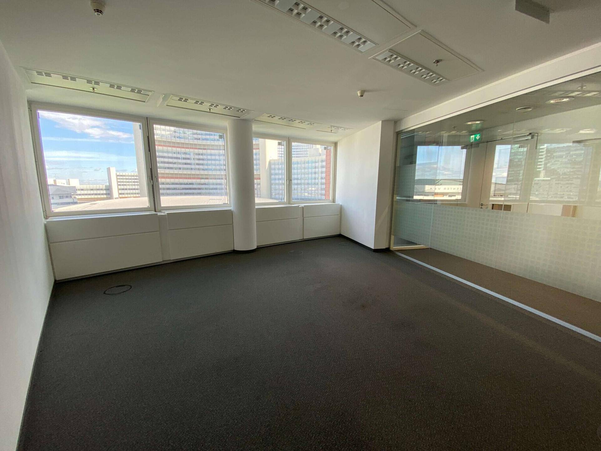 Einzelbüro mit Teppichboden und großen Fenstern, die einen Blick auf die umliegenden Gebäude ermöglichen.