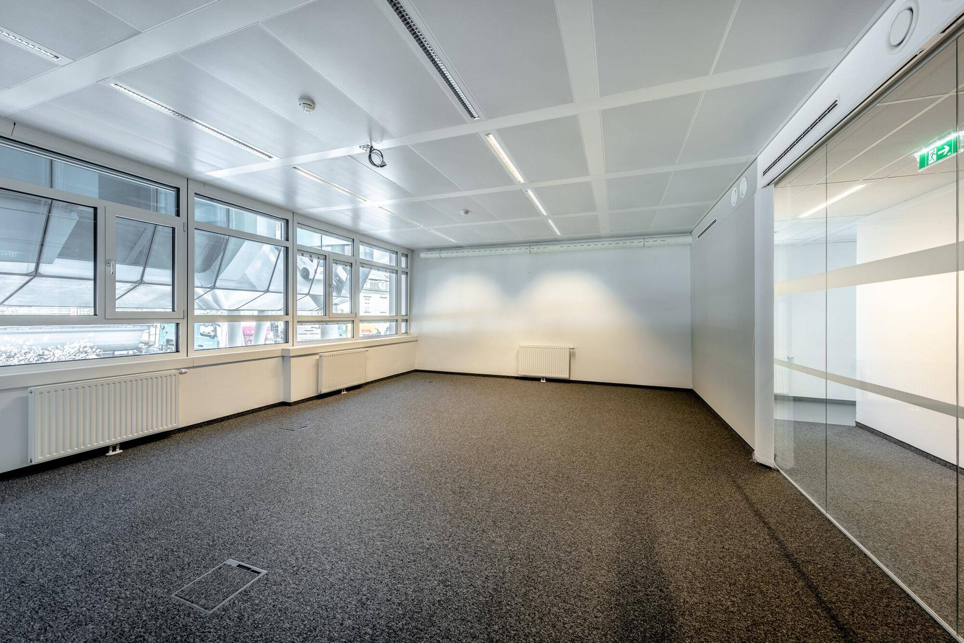 Leerer Büroraum mit Teppichboden, großen Fenstern und einer Glaswand, die Tageslicht hereinlässt.