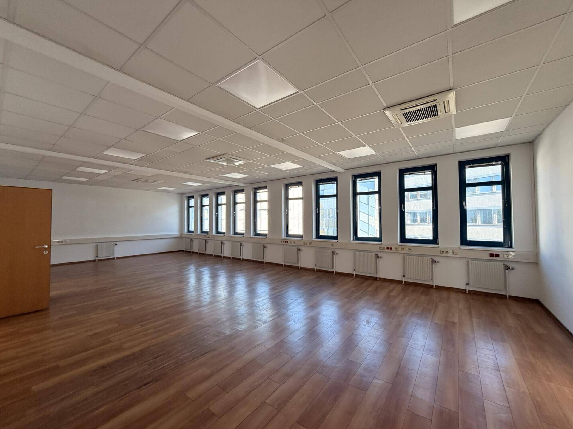 Geräumiger, leerer Büroraum mit Holzboden, weißen Wänden und vielen Fenstern.