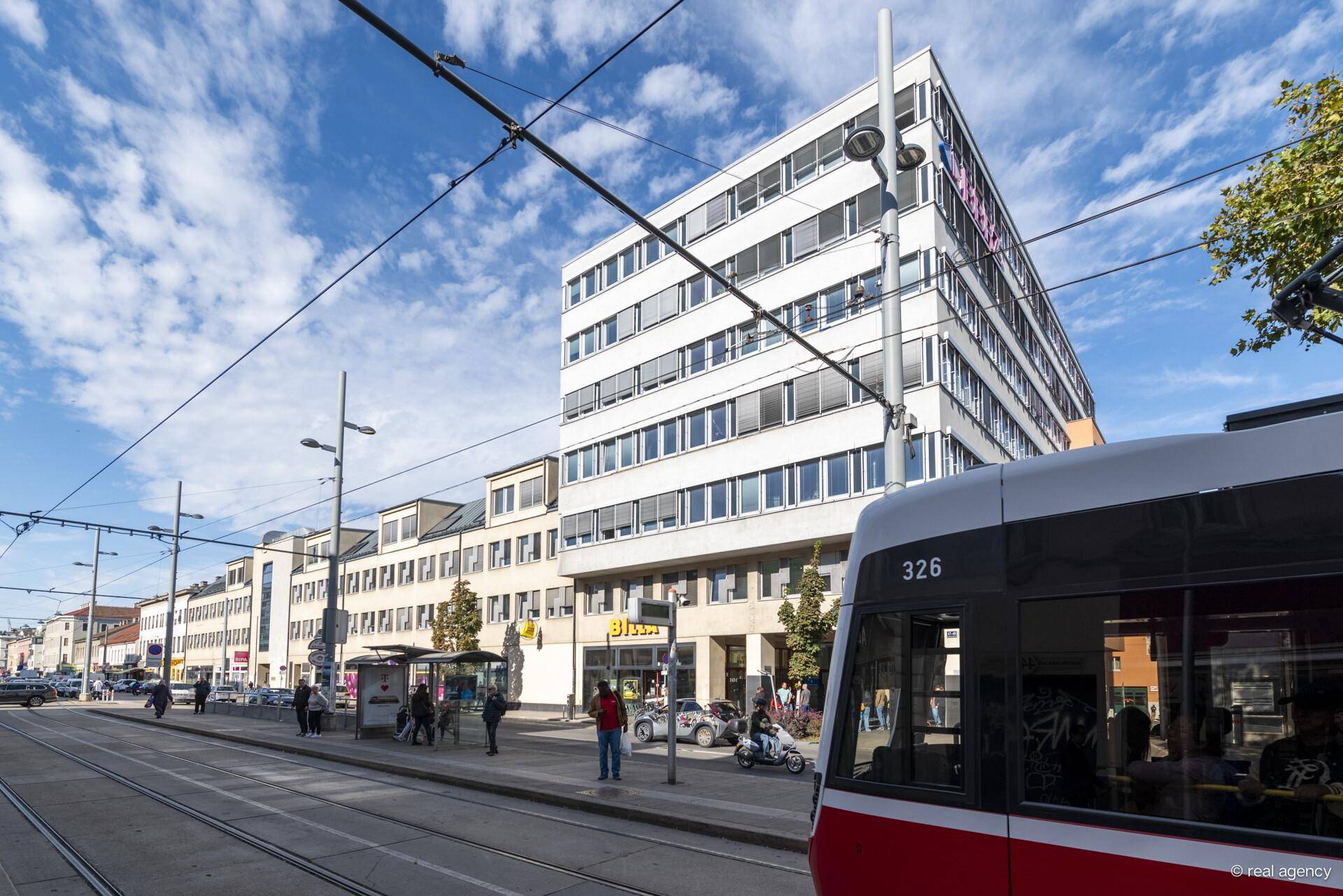 Bürogebäude an einer Straße mit Straßenbahn, Geschäften im Erdgeschoss und blauem Himmel.