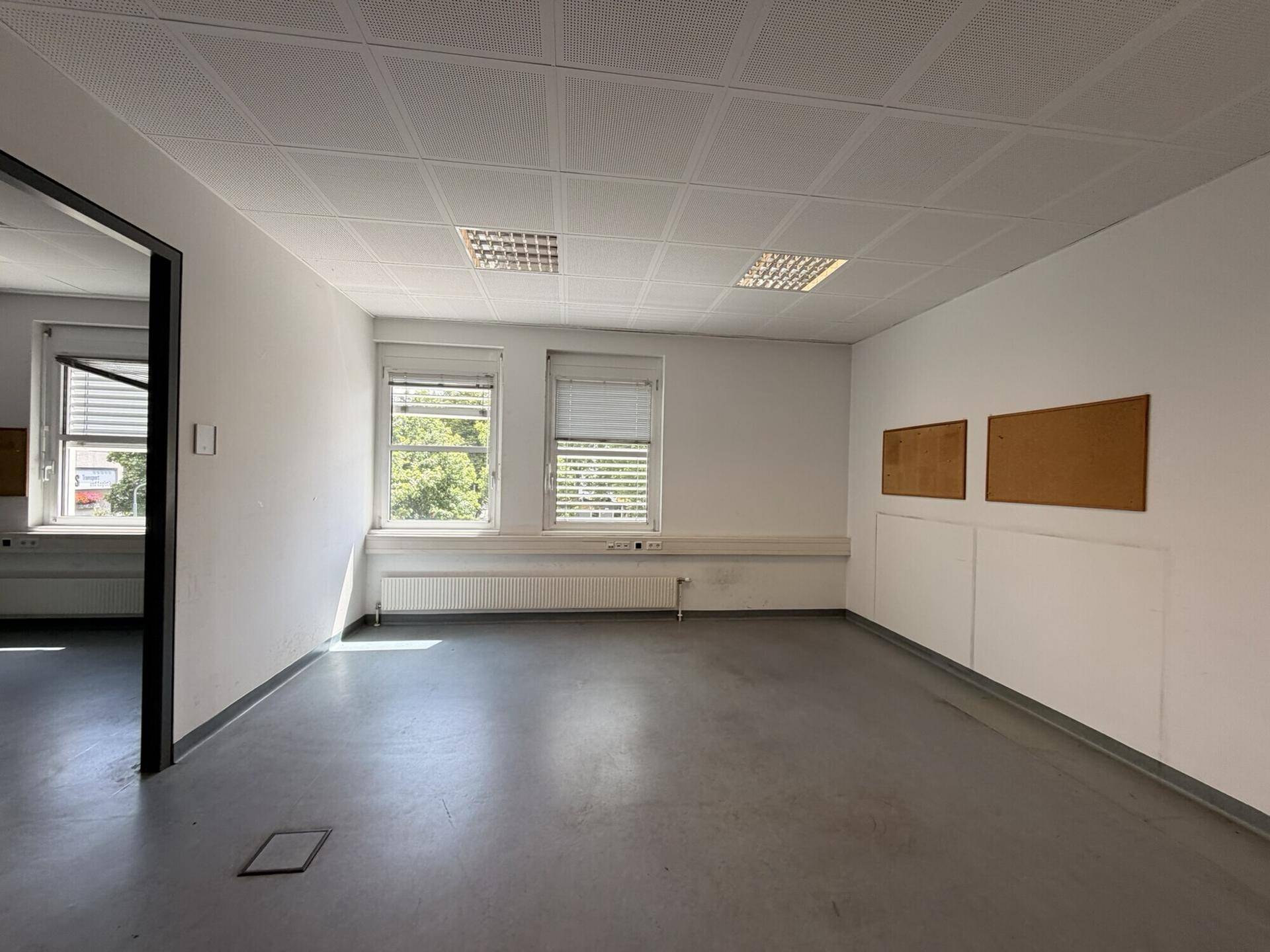 Heller Büroraum mit zwei Fenstern, Pinnwänden und einem grauen Bodenbelag, ideal für kreative Arbeit.