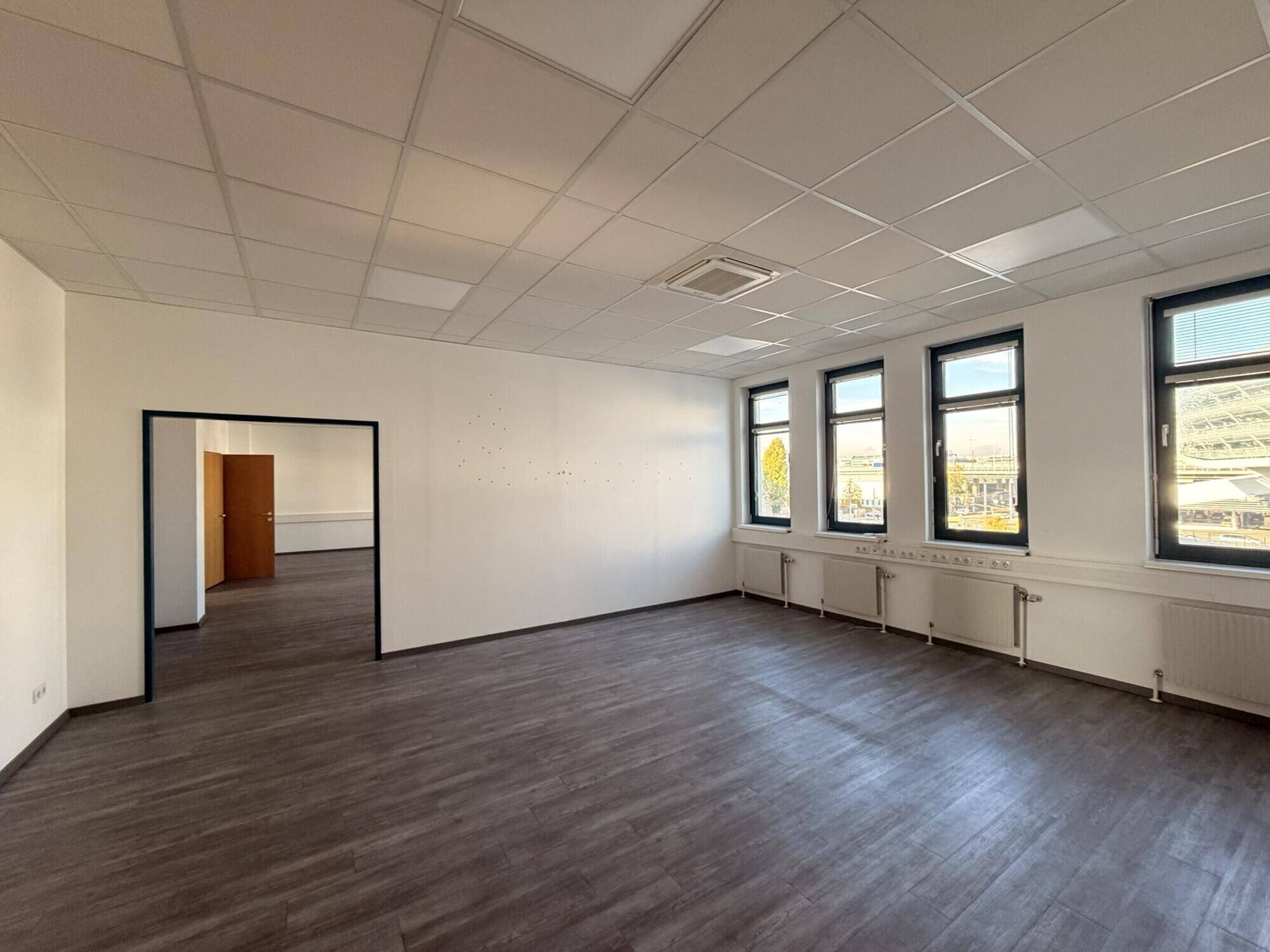 Geräumiger Büroraum mit dunklem Laminatboden und vielen Fenstern für Tageslicht.