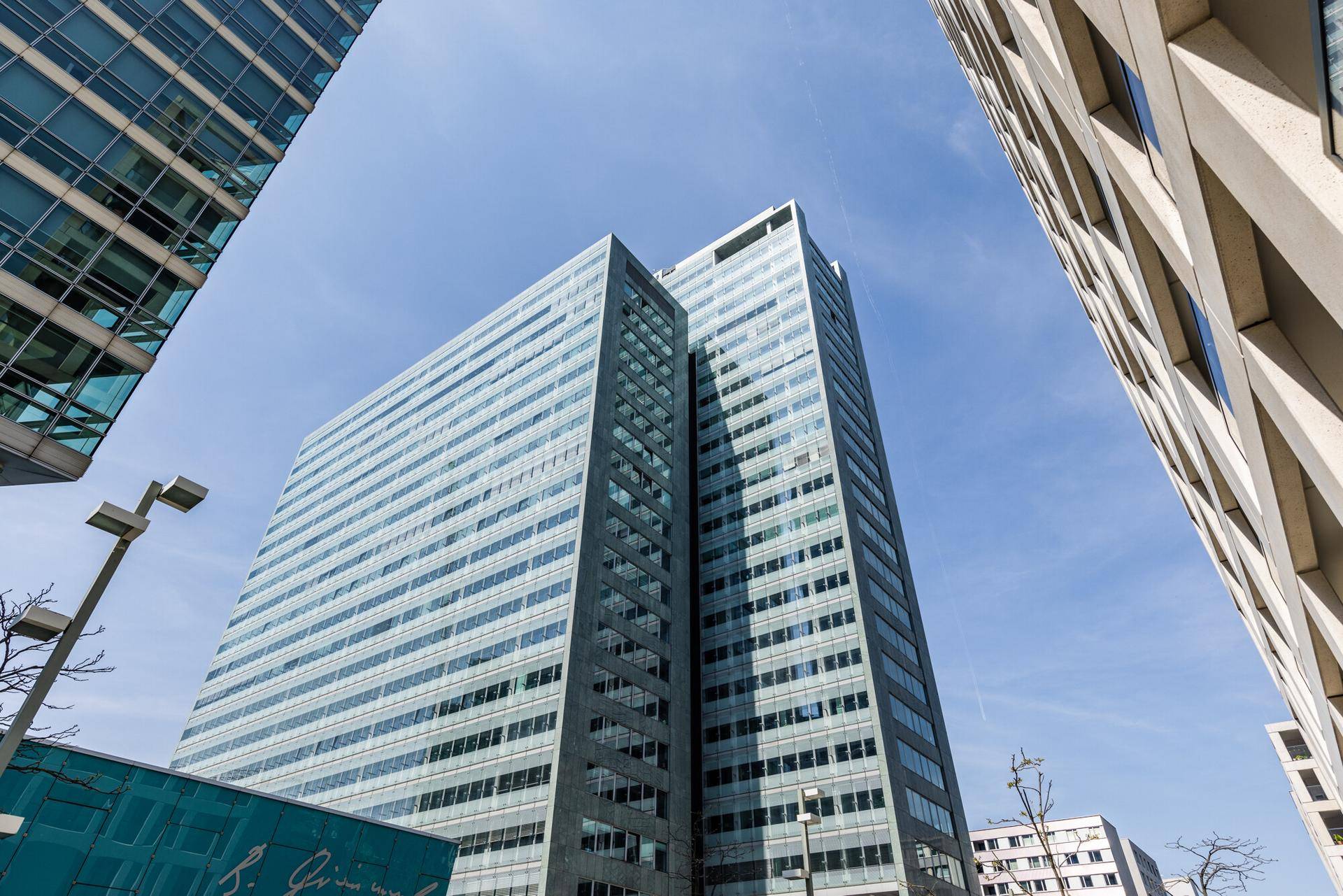 Moderne Hochhausfassade mit vielen Glasflächen unter blauem Himmel, umgeben von anderen Gebäuden.