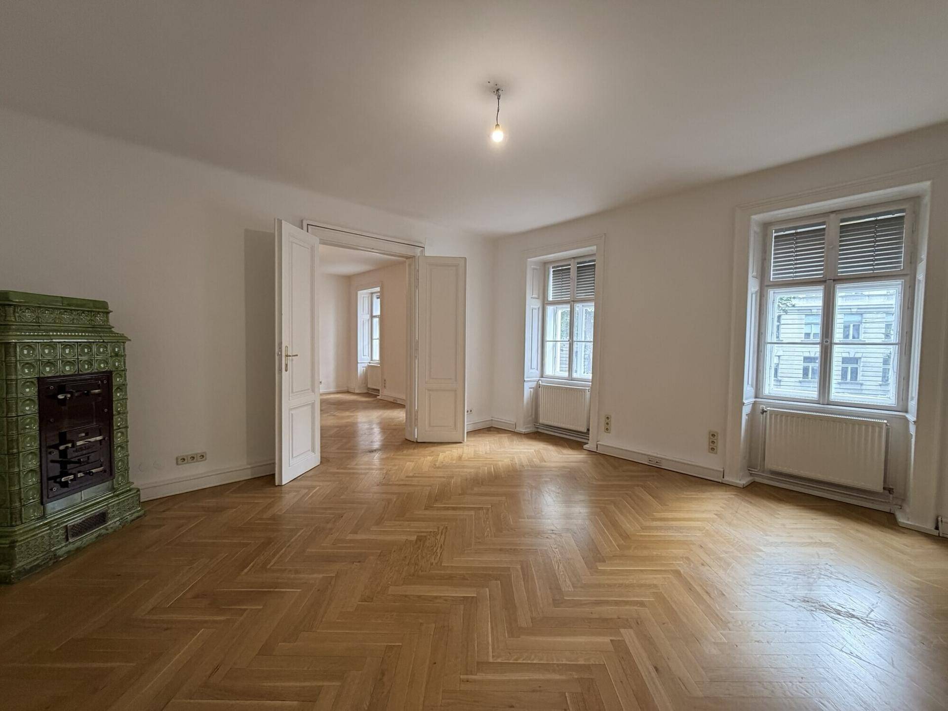 Geräumiges Wohnzimmer mit Fischgrätparkett, hohen Fenstern und einem traditionellen Kachelofen.