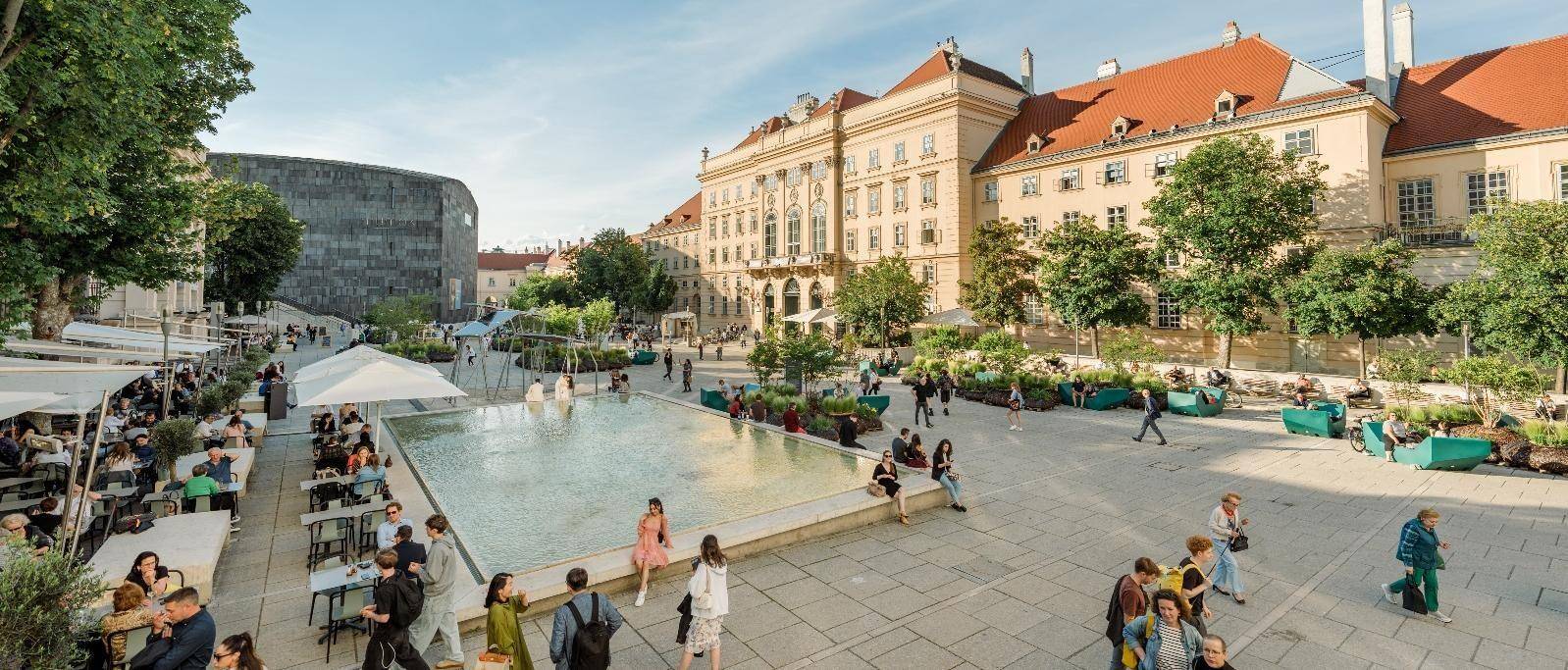Belebtes Stadtbild mit historischer Architektur, einem modernen Gebäude und einem öffentlichen Platz mit Wasserbecken.