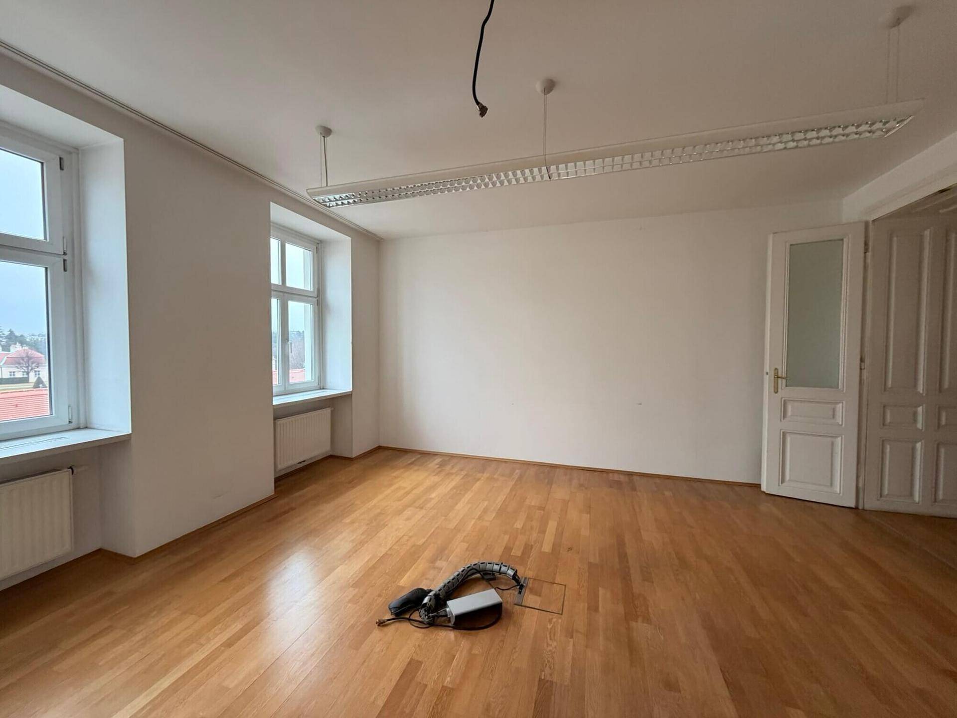 Leerer Büroraum mit Holzboden, großen Fenstern und einer Deckenleuchte, bereit zur individuellen Einrichtung und Nutzung.