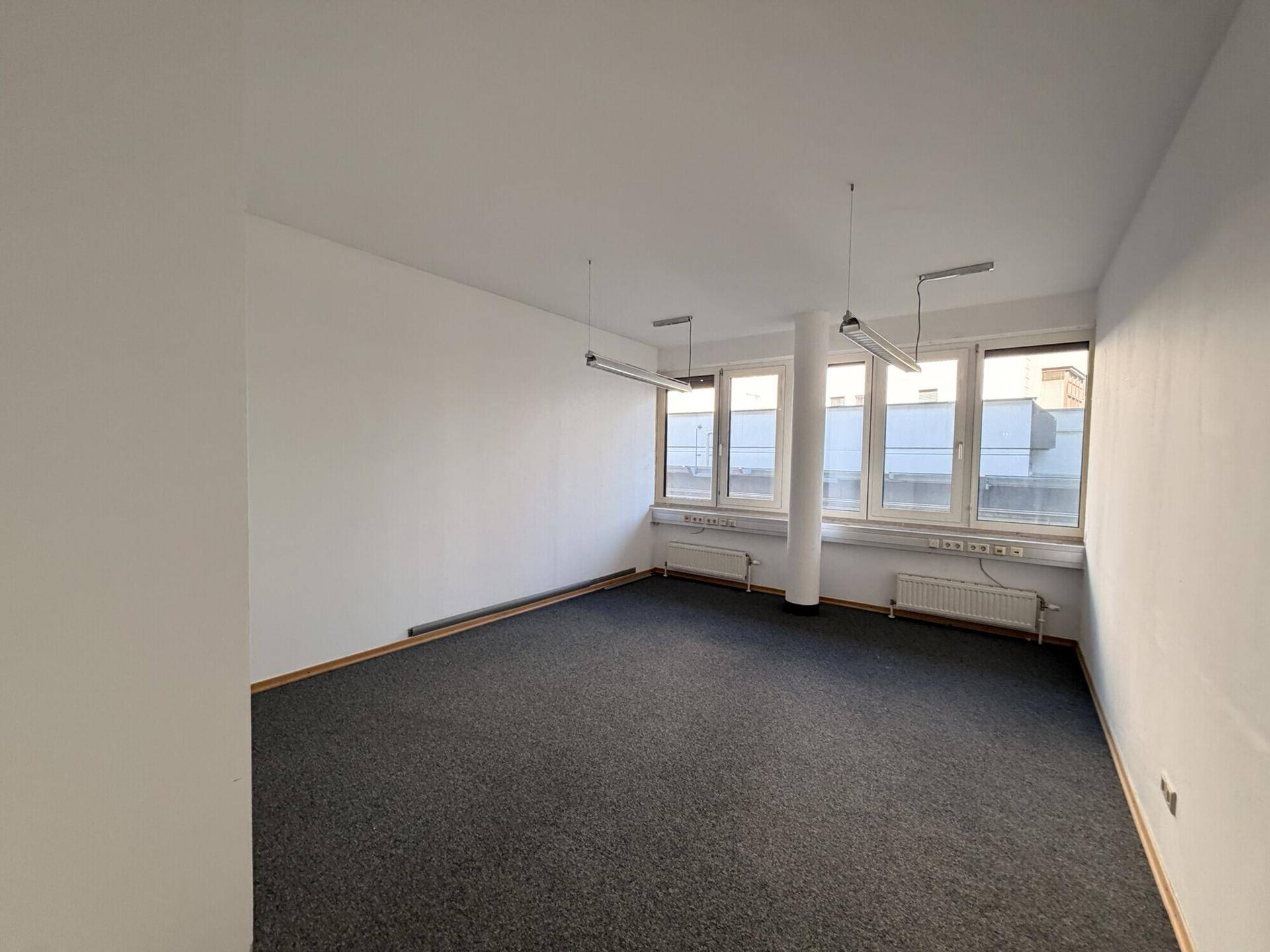 Geräumiges, leeres Büro mit Teppichboden, Säule und Fenstern, die für natürliche Beleuchtung sorgen.