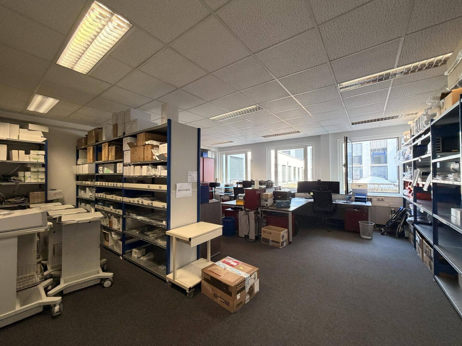 Geräumiges Büro mit Regalen für Lagerung und mehreren Arbeitsplätzen, gut beleuchtet.