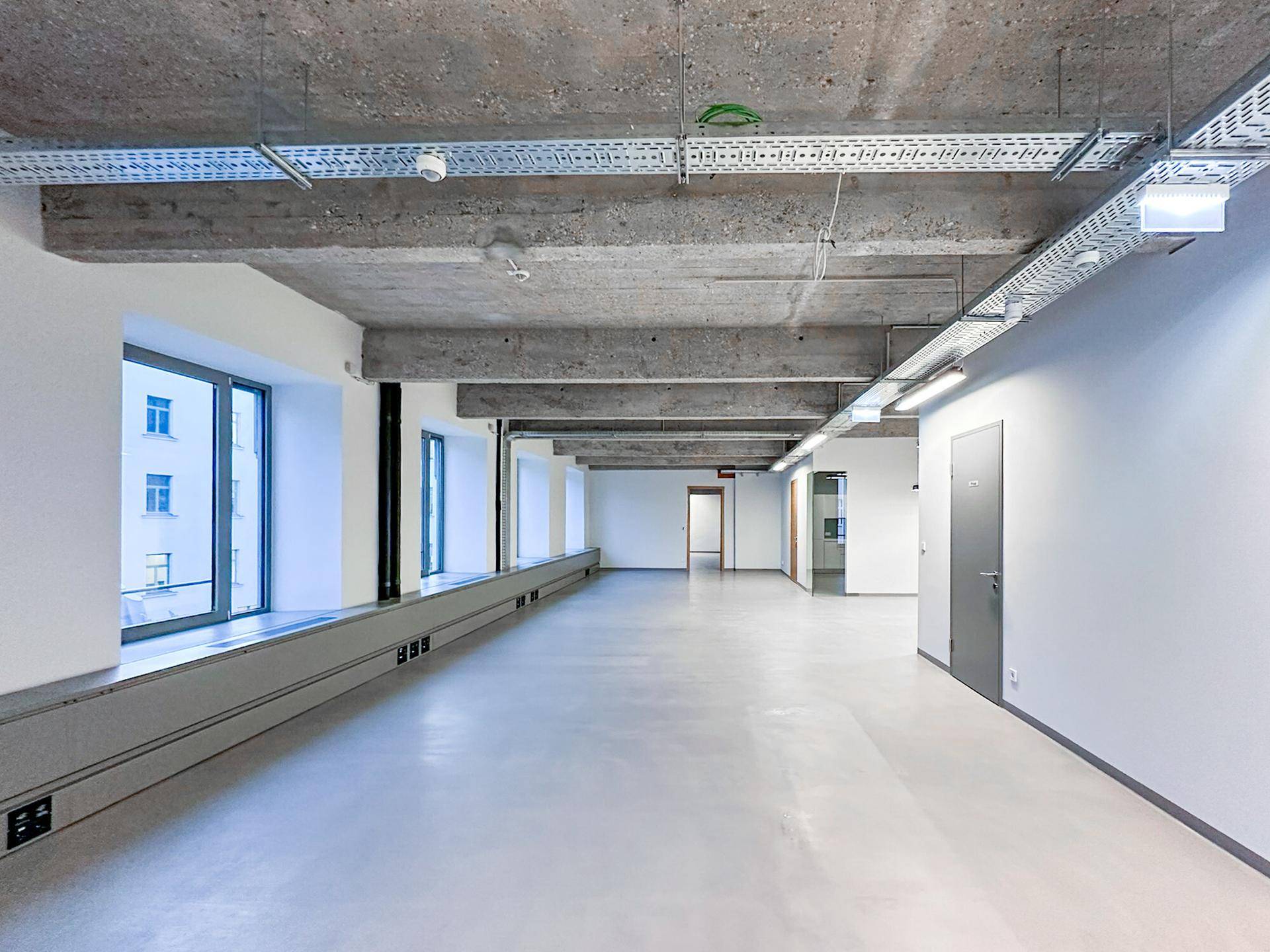 Geräumiger, leerer Büroraum mit Sichtbetondecke, großen Fenstern und freiliegenden Installationen im Industriestil.