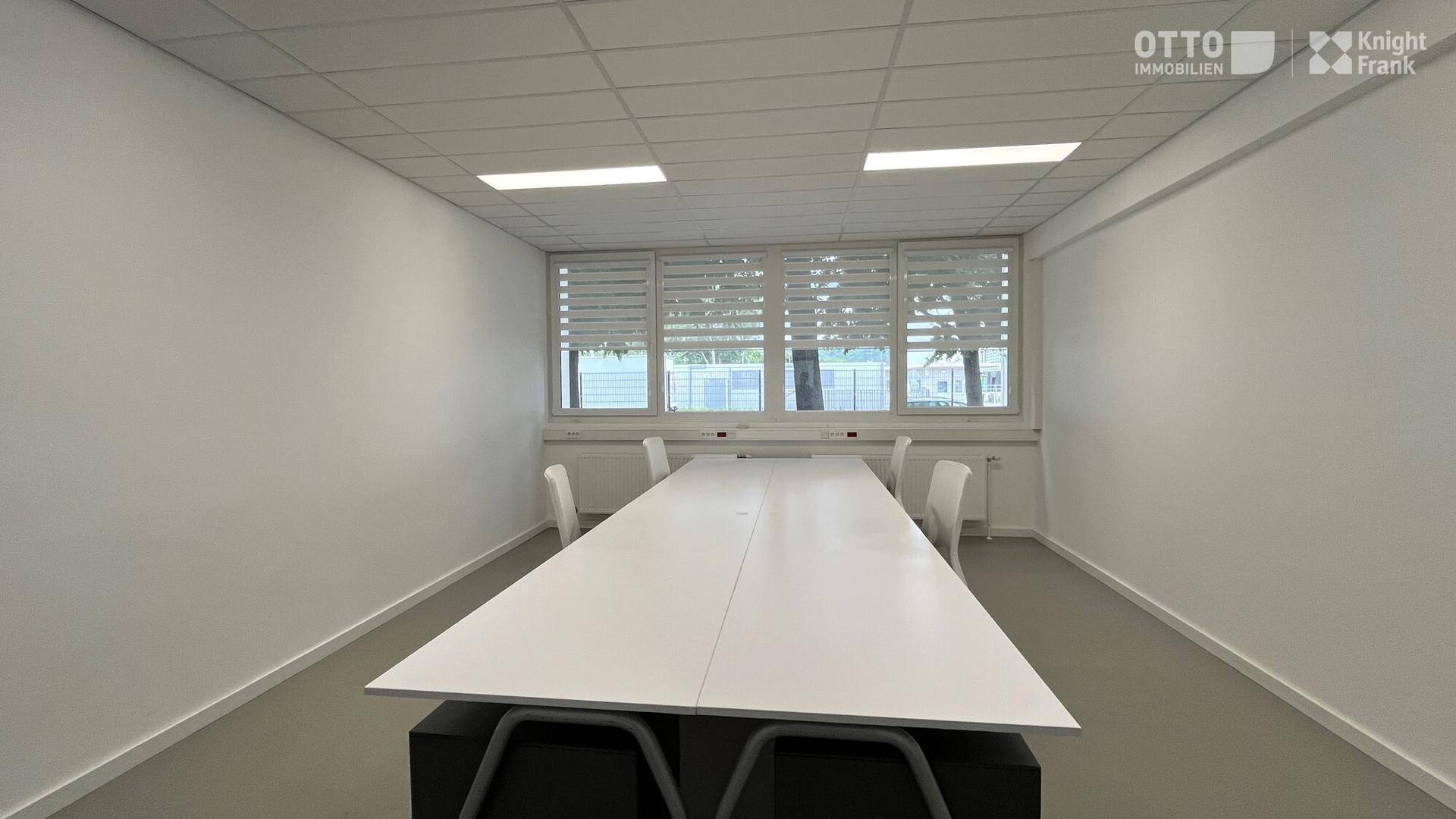 Moderner Konferenzraum mit einem großen weißen Tisch und Stühlen, beleuchtet durch Fenster mit Jalousien.