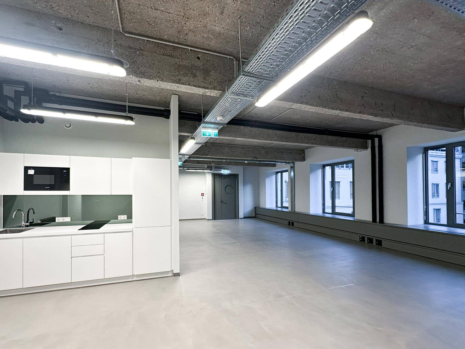 Moderner Büroraum mit integrierter Teeküche, Sichtbetondecke und großen Fenstern für viel Licht.