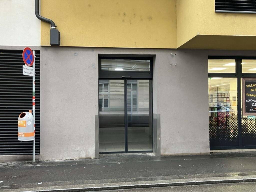 Moderner Gebäudeeingang mit automatischen Glastüren und einer schlichten grauen Fassade an einer städtischen Straße.