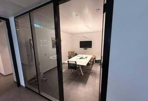 Moderner Besprechungsraum mit Glastür, großem Tisch und Wandfernseher, ideal für kleine Meetings.