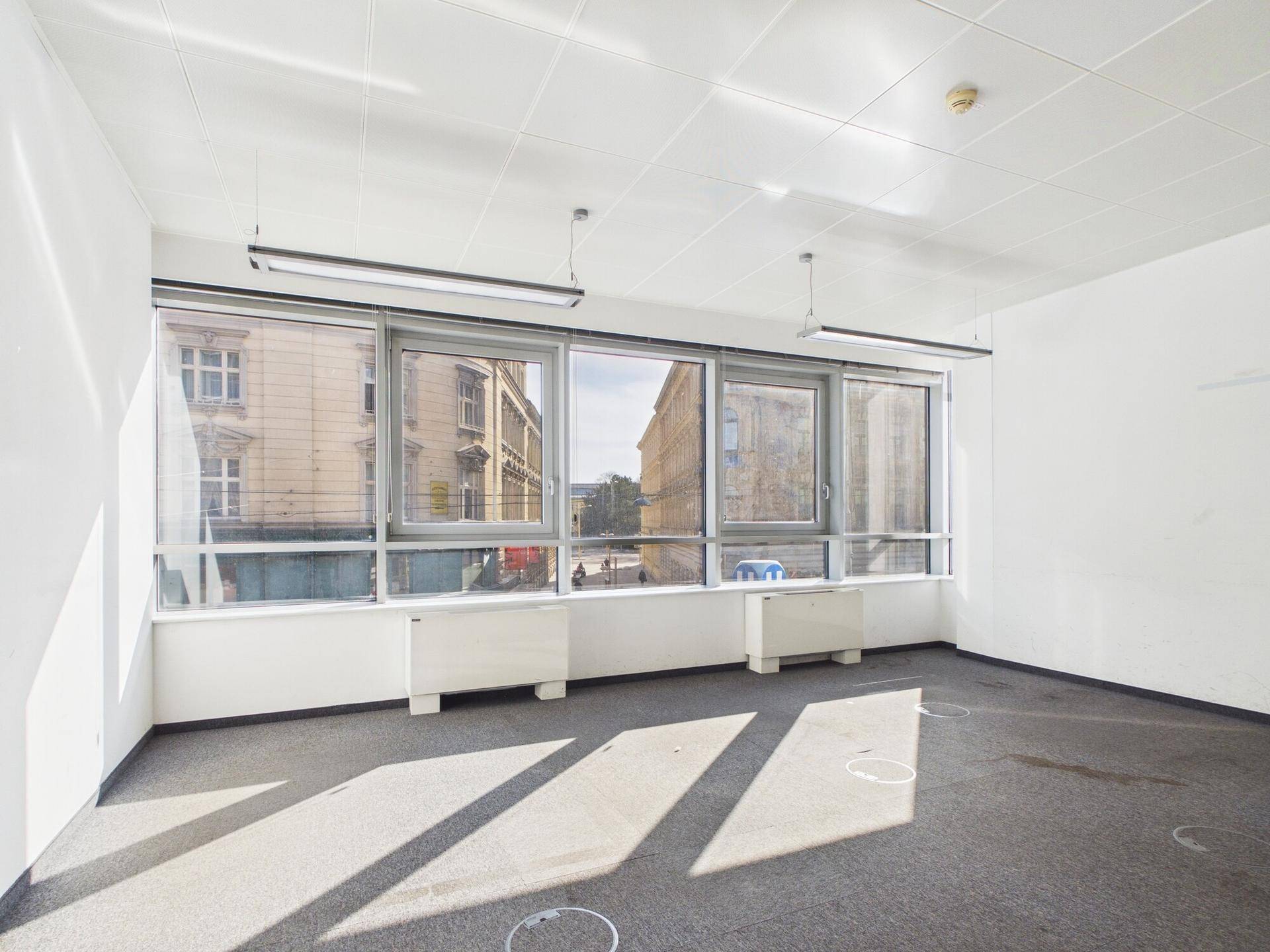 Helles Büro mit Teppichboden und breiter Fensterfront, die einen Blick auf die umliegenden Gebäude ermöglicht.