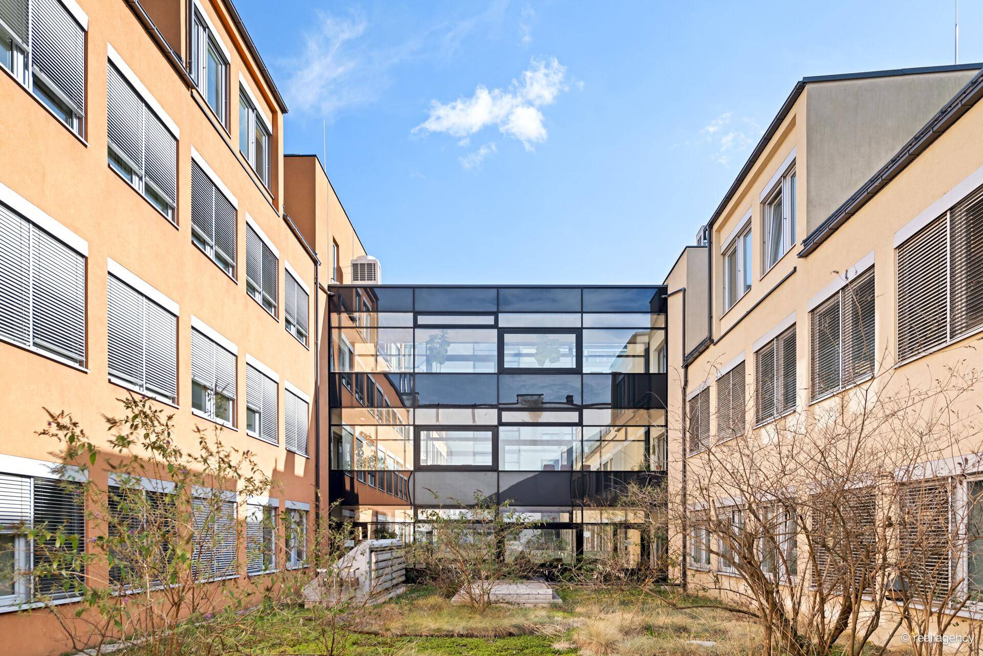 Innenhof zwischen Gebäuden mit einer modernen Glasbrücke und natürlicher Bepflanzung.