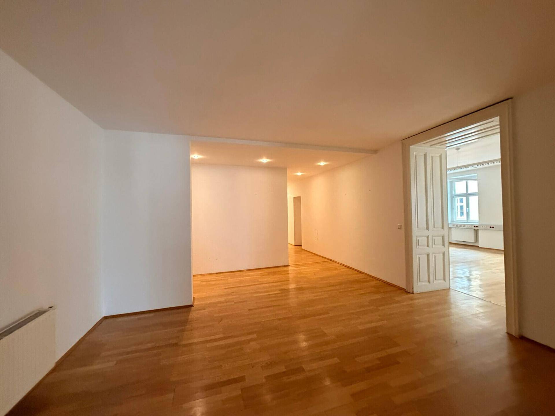 Geräumiger, leerer Büroraum mit Holzboden und weiß gestrichenen Wänden, der in weitere Räume führt und viel Platz bietet.