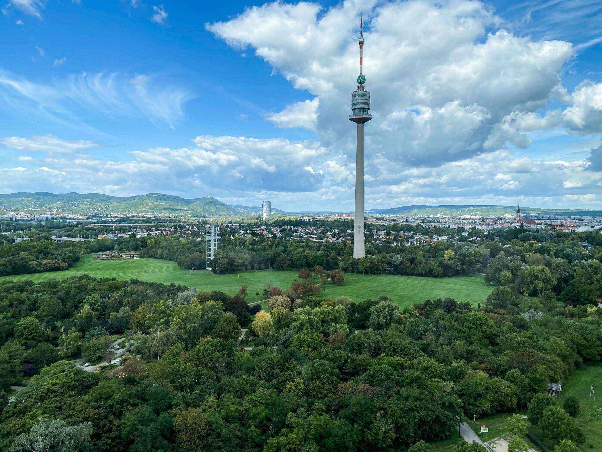 Panoramablick auf die grüne Umgebung mit dem Donauturm und der Stadt im Hintergrund unter blauem Himmel.