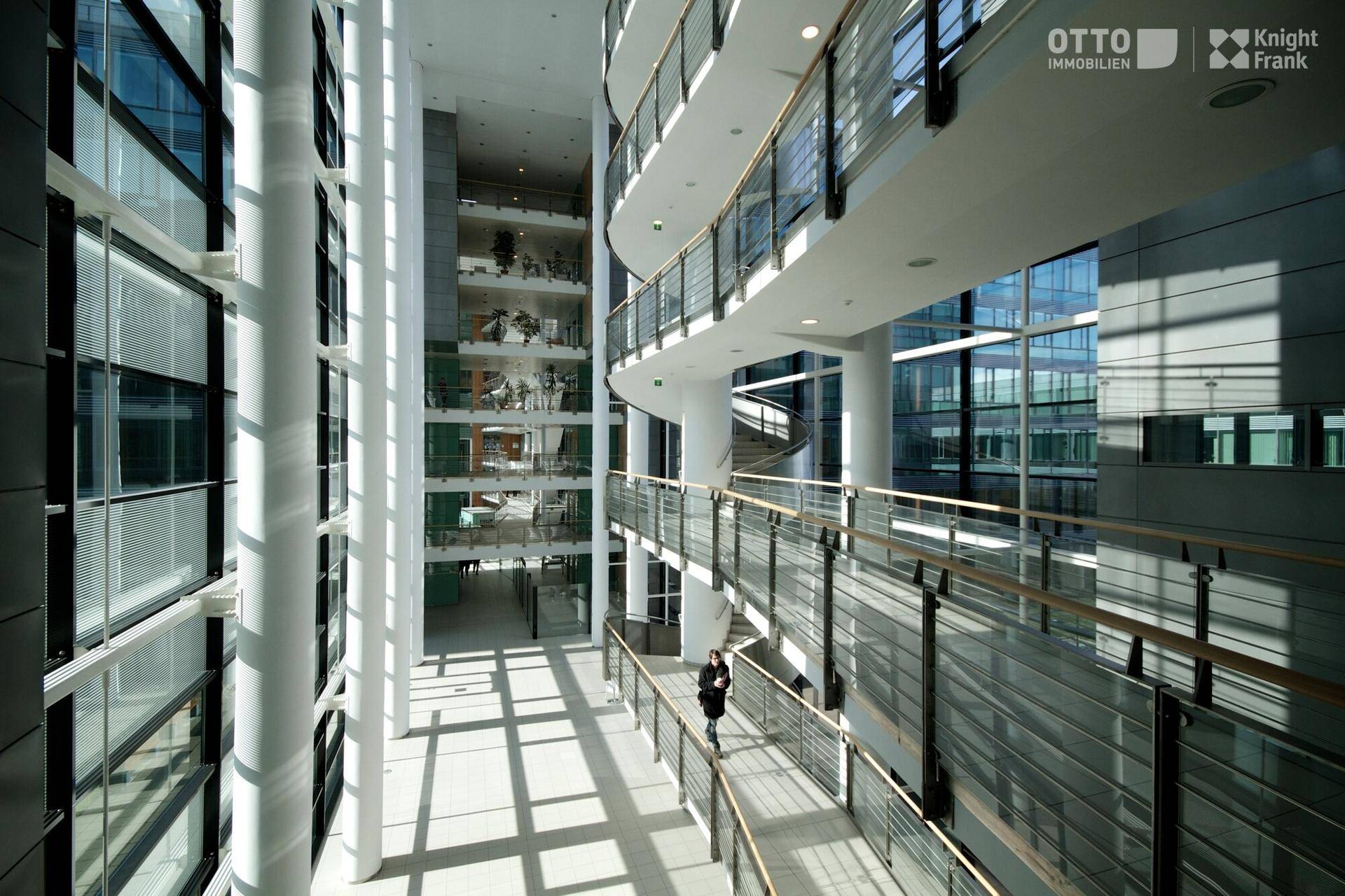 Helles, modernes Atrium mit mehreren Etagen, Glasgeländern und einer eleganten Wendeltreppe im Hintergrund.