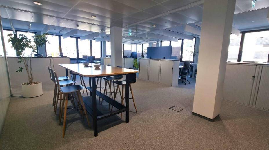 Großes, offenes Büro mit Teppichboden, hohen Tischen und vielen Fenstern, die eine helle Arbeitsumgebung schaffen.