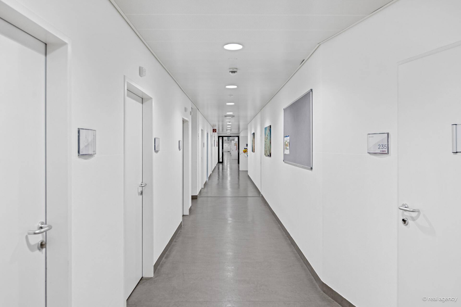 Langer, sauberer Flur mit weißen Wänden, vielen Türen und einem grauen Boden.