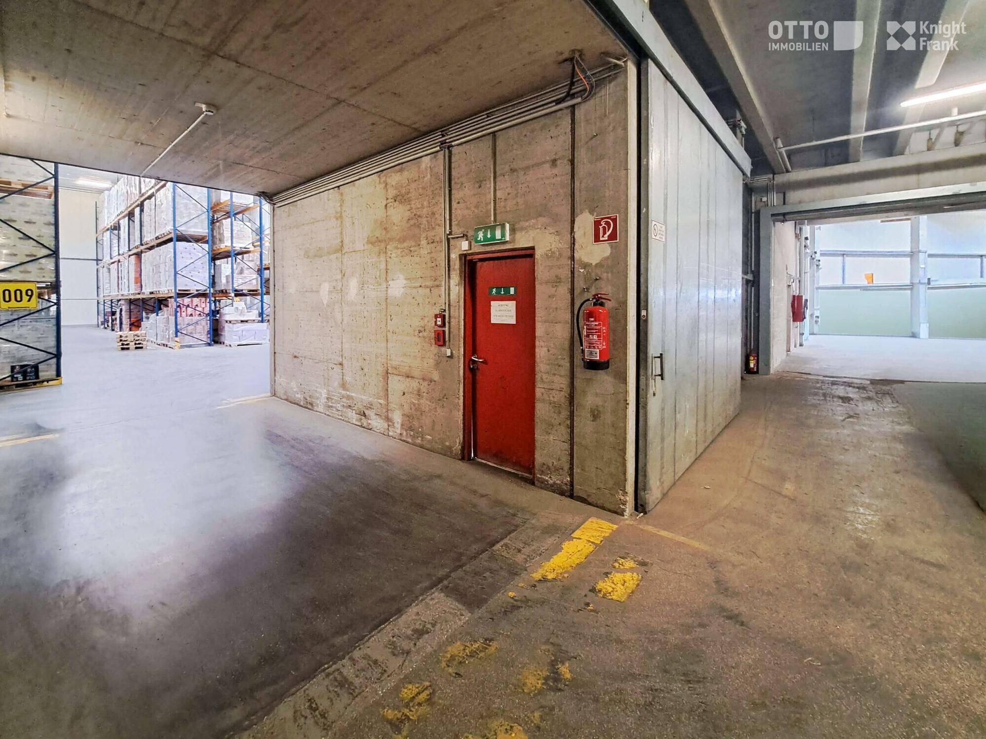 Eingangsbereich einer Lagerhalle mit Betonwänden, roter Tür und Feuerlöscher, führt zu den Lagerbereichen.
