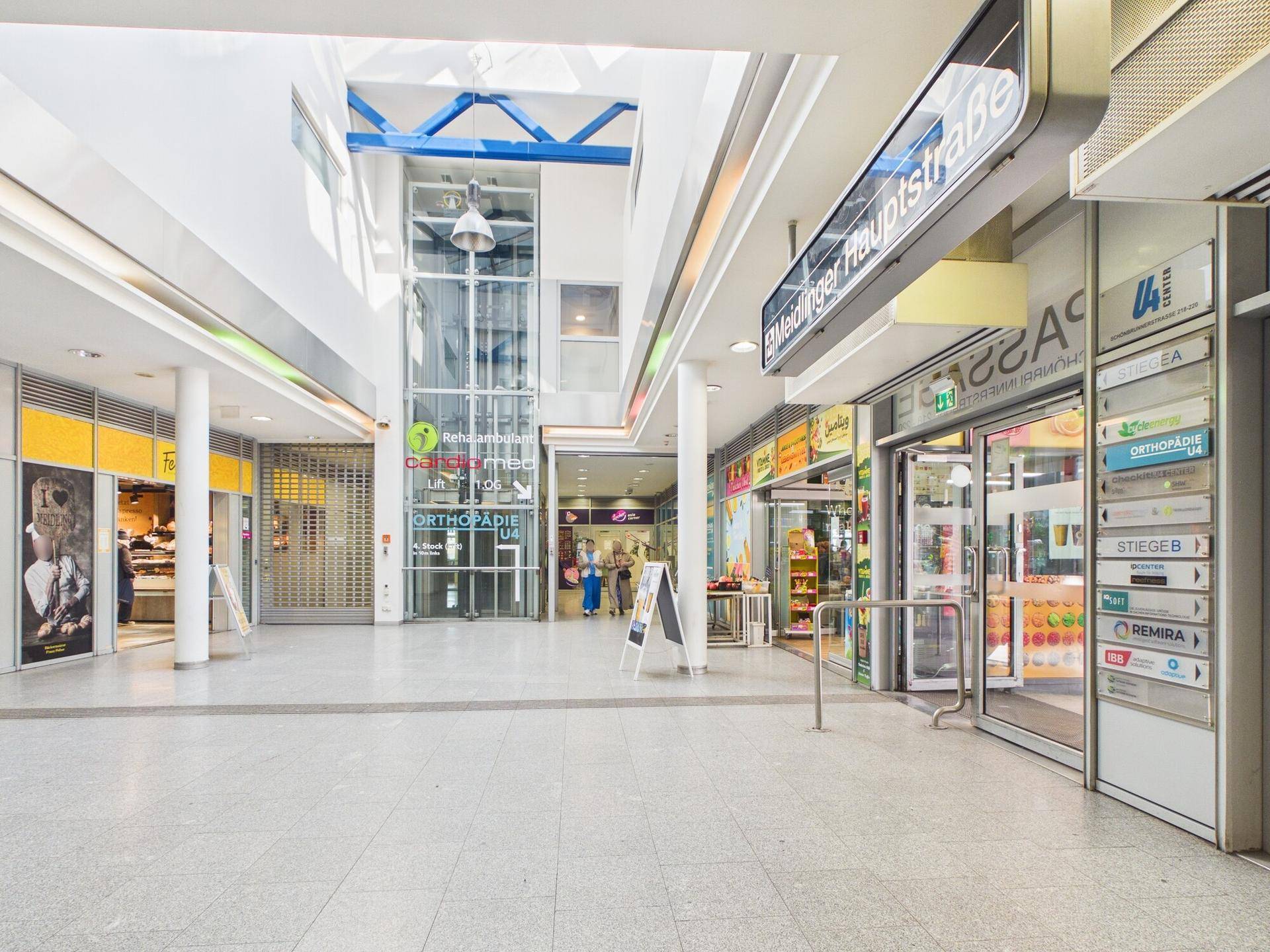 Helle Ladenpassage mit verschiedenen Geschäften und einem gläsernen Aufzug in der Mitte.
