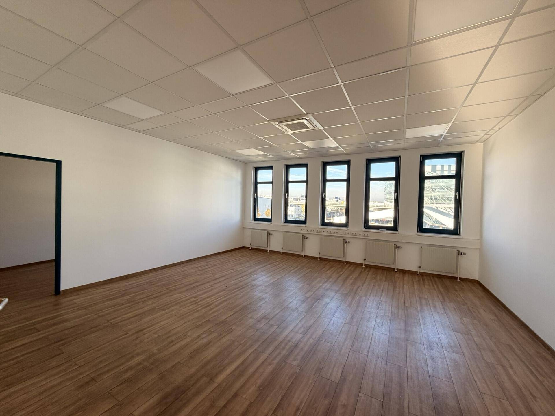 Leerer Büroraum mit hellem Holzboden, weißen Wänden und Fenstern, die für viel Tageslicht sorgen.