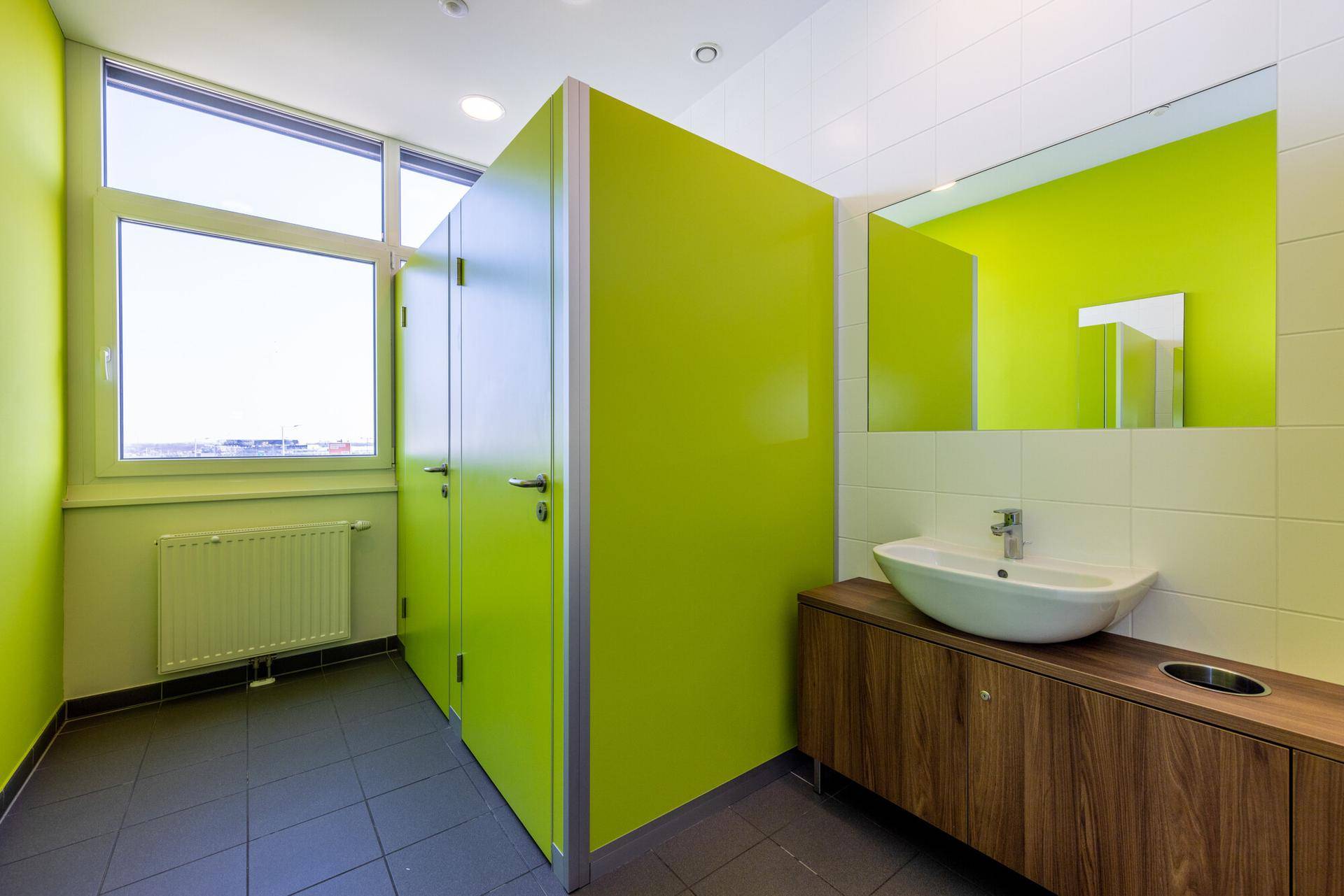 Moderne Toilettenanlage mit leuchtend grünen Kabinen, Fliesenboden und einem Fenster für Tageslicht.