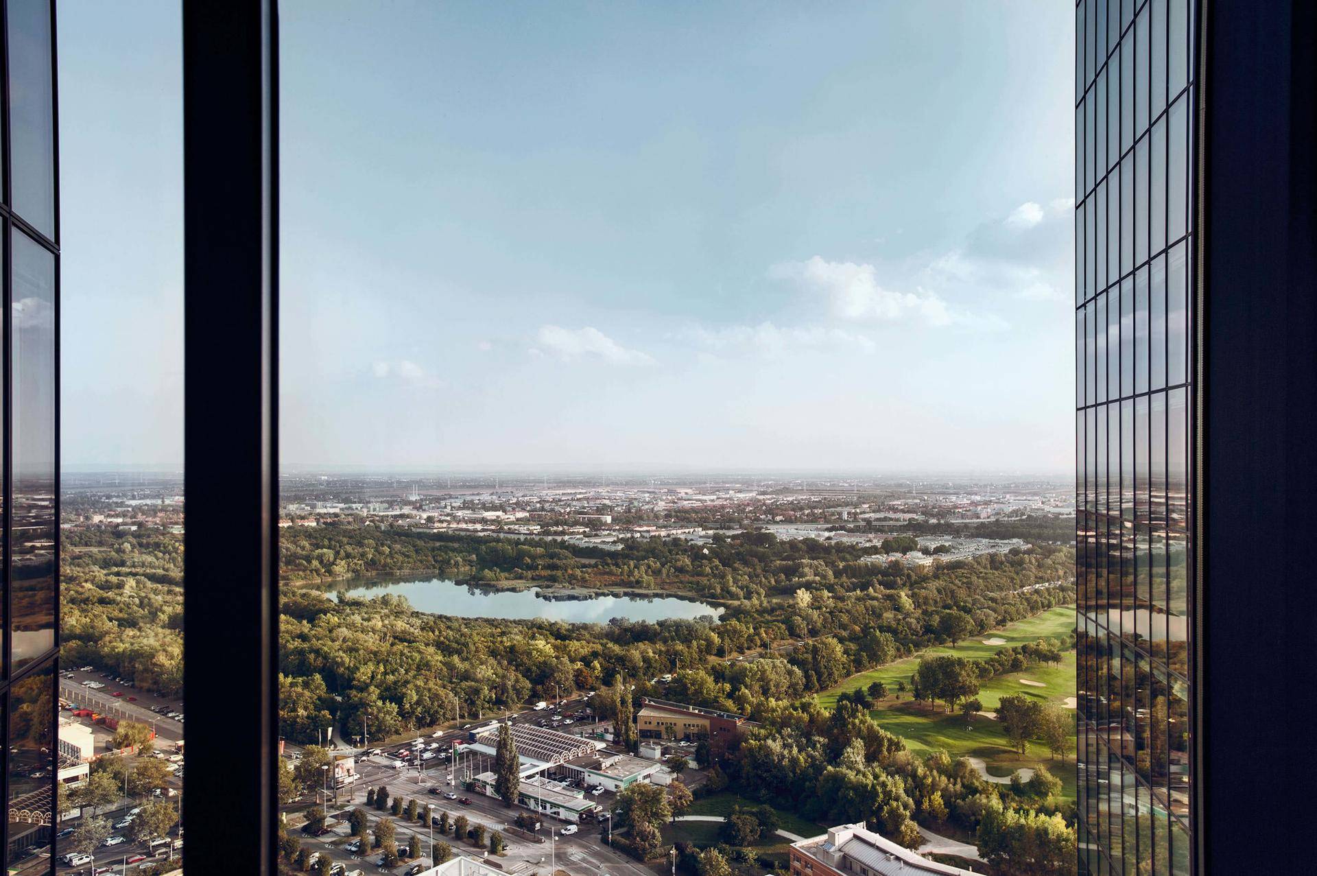 Panoramablick aus einem Hochhausfenster auf einen See, weitläufige Grünflächen und die umliegende Stadtlandschaft.