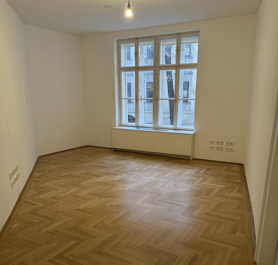 Helles, leeres Zimmer mit großen Fenstern und klassischem Fischgrätparkett, vielseitig nutzbar als Büro oder Wohnraum.
