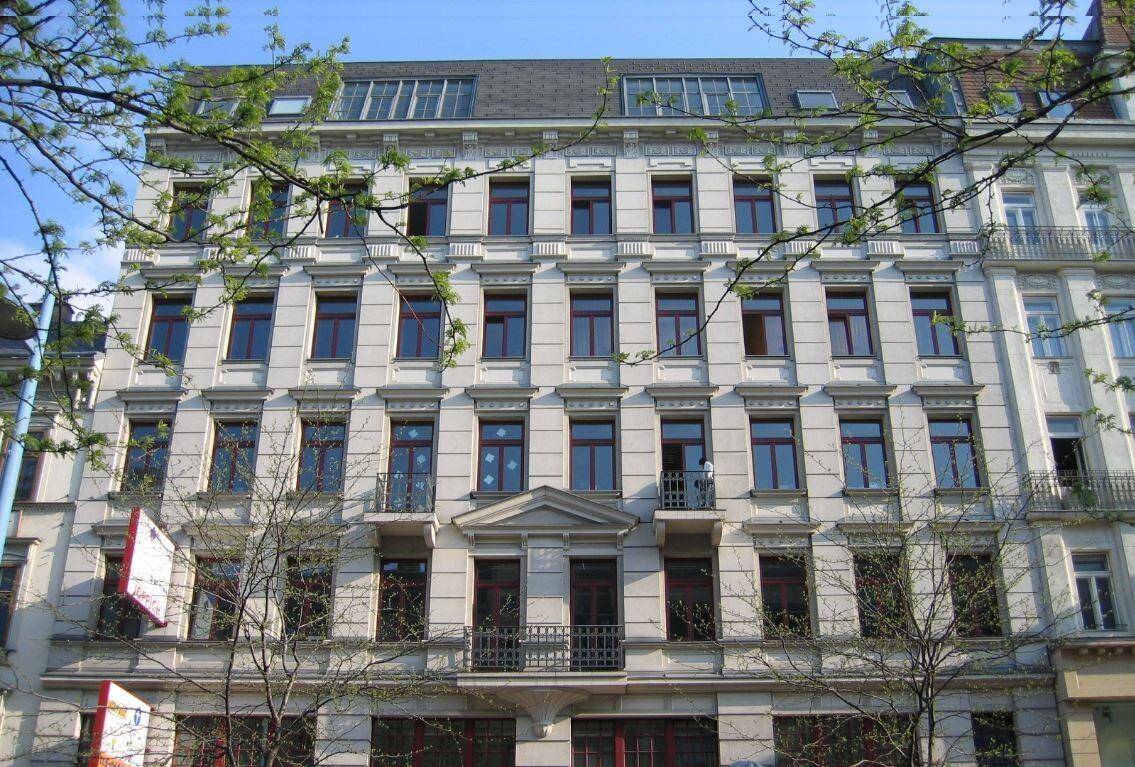 Klassische Altbaufassade mit vielen Fenstern und dekorativen Elementen, umgeben von Bäumen.
