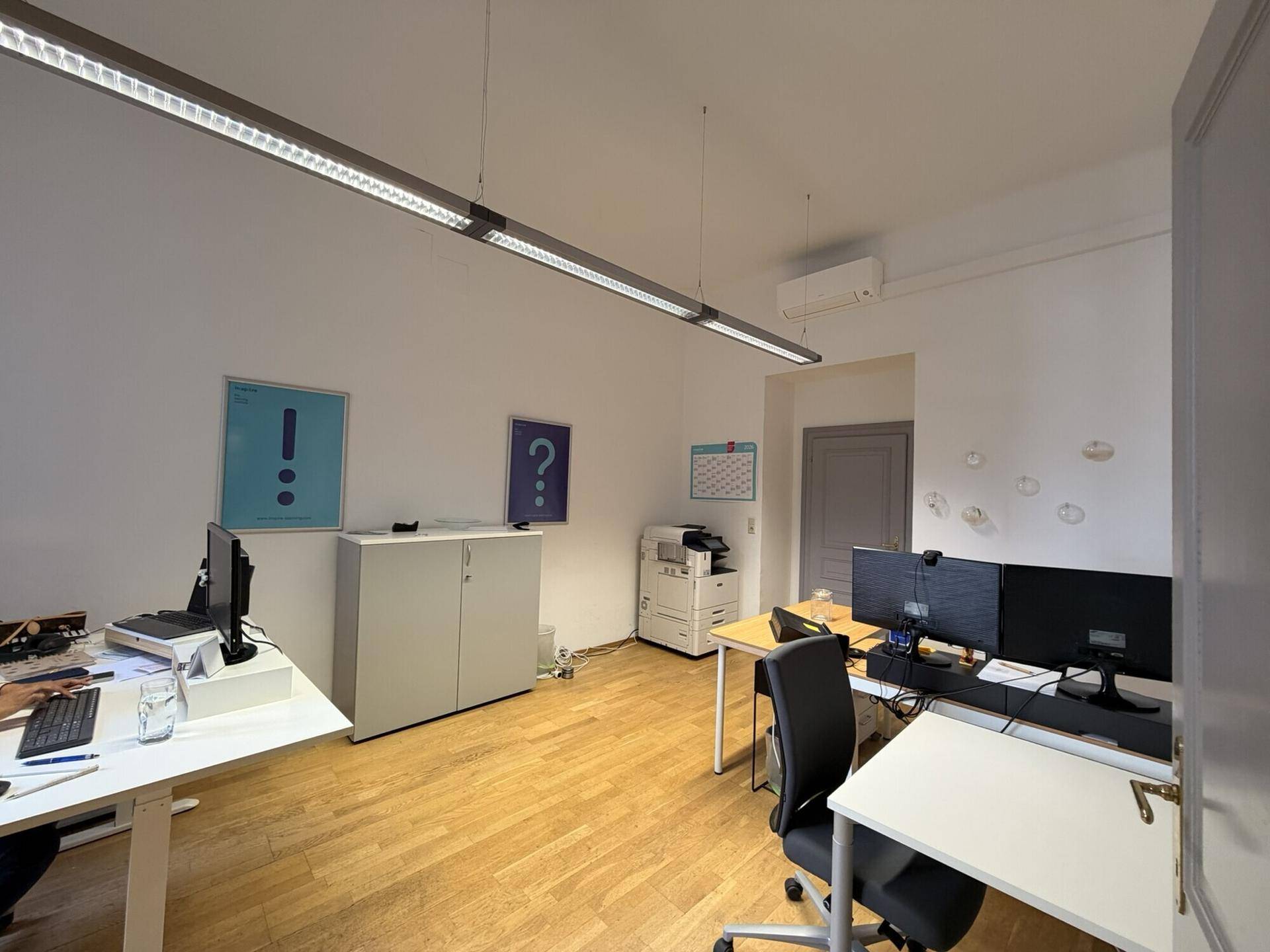 Funktionales Büro mit mehreren Arbeitsplätzen, Holzboden und einem Klimagerät an der Wand.