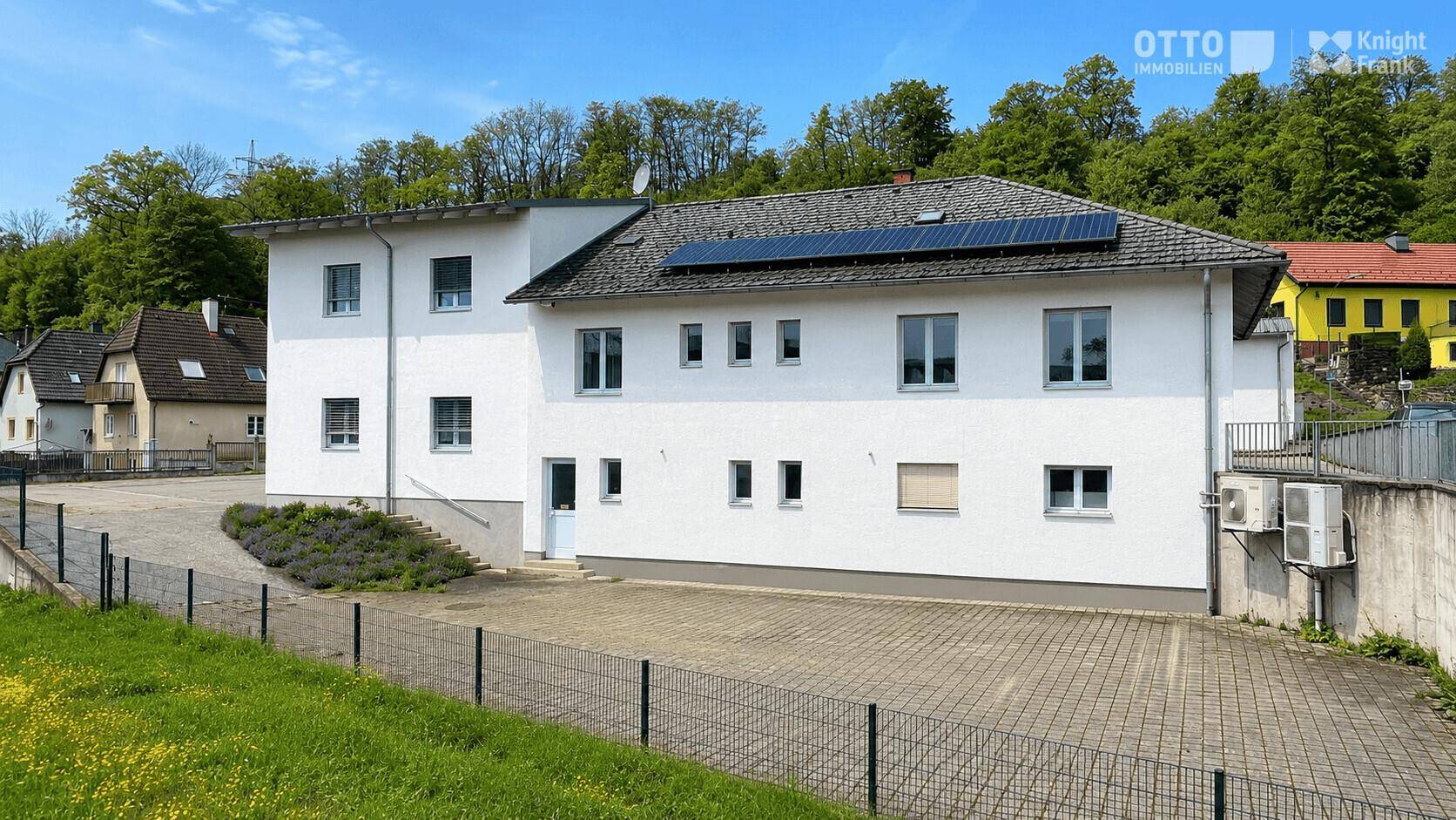 Seitenansicht eines weißen Gebäudes mit Solarpaneelen auf dem Dach, gepflastertem Bereich und grüner Umgebung.