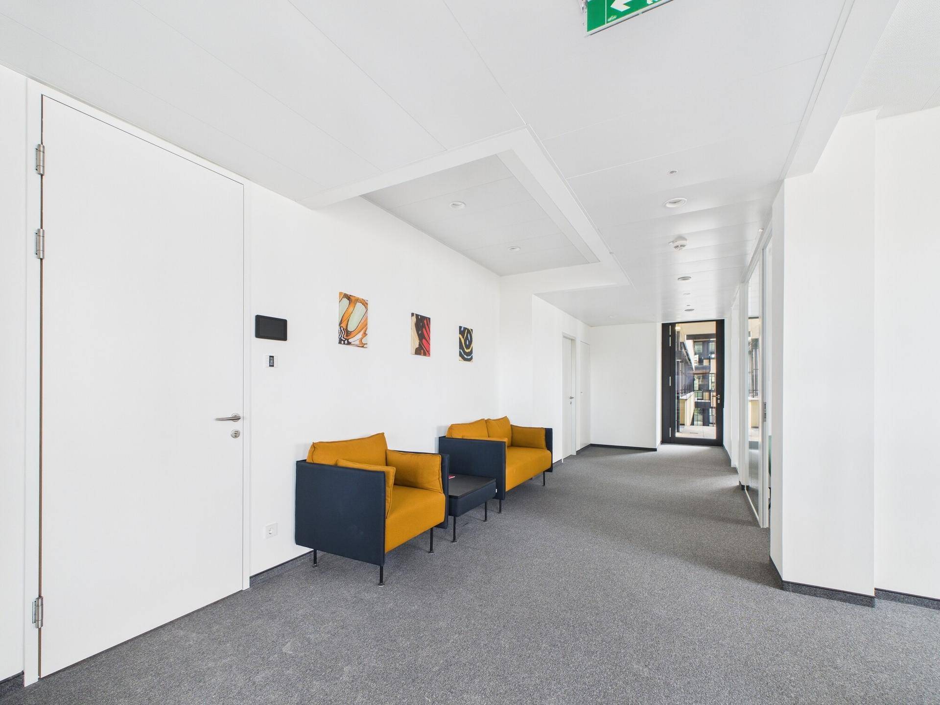 Heller Bürogang mit modernen Sitzgelegenheiten, Kunstwerken und Zugang zu weiteren Räumen, einladend gestaltet.